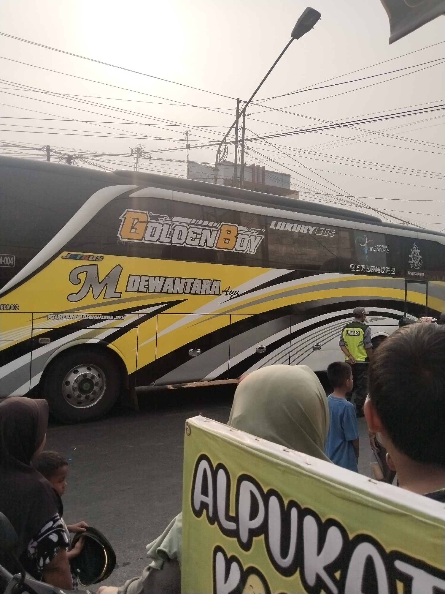 In Balapulang Kulon, Indonesia, a parked luxury bus, identified by "GOLDEN BOY" and "JETBUS" branding, is visible on a street. The bus features a yellow and black livery with white accents and the text "M DEWANTARA Ayu". A man in a yellow vest with "LINMAS" written on the back stands near the bus. Several people are in the foreground, including a woman wearing a hijab and a child holding a hat, partially obscuring a yellow banner with the text "ALPULKA". The background shows a hazy sky filled with a dense network of overhead electrical wires and poles, suggesting an urban or semi-urban environment.