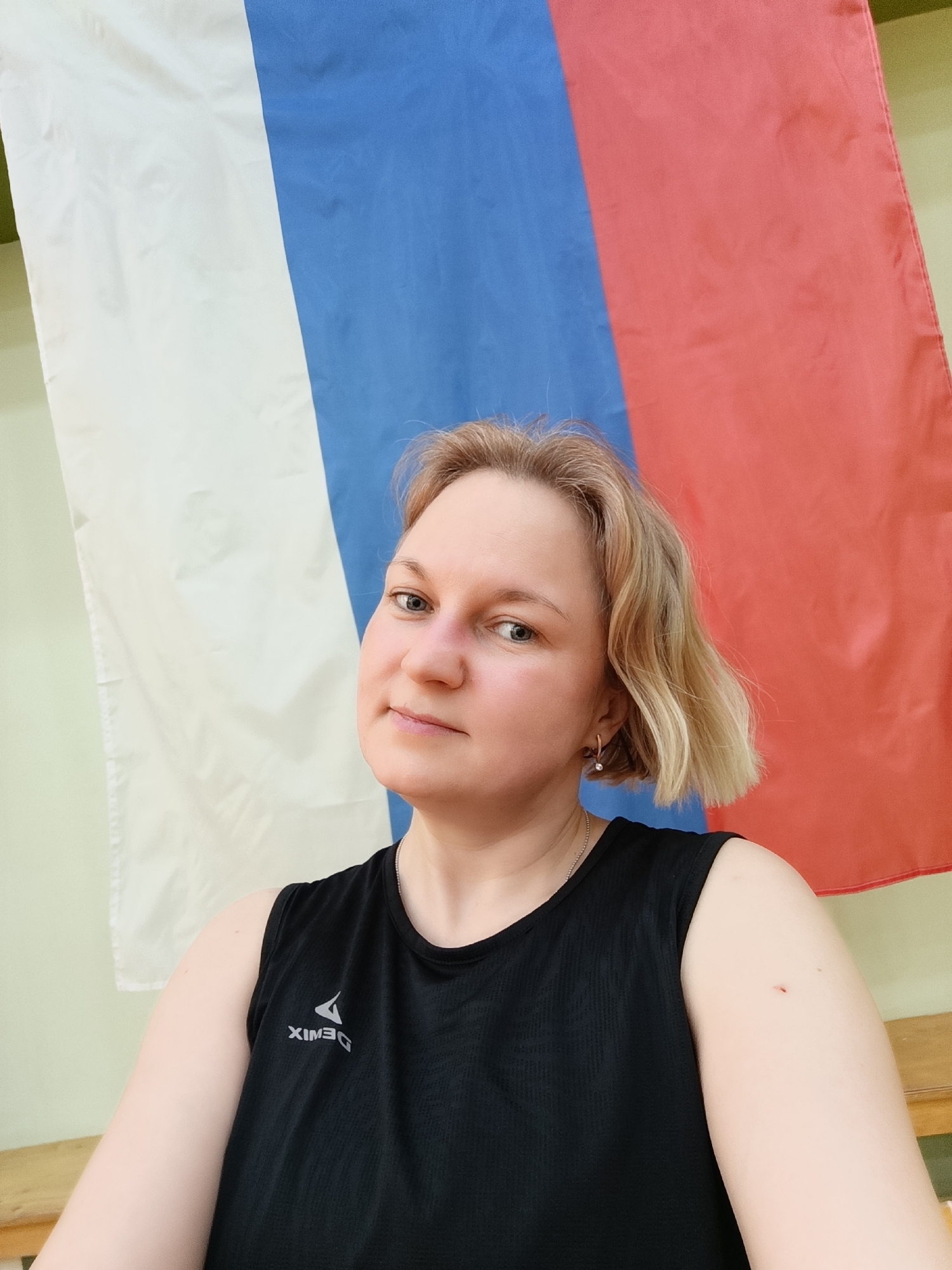 A medium close-up portrait shows a woman with shoulder-length, wavy blonde hair and fair skin, looking directly at the camera with a neutral expression. She has light-colored eyes and a hint of a slight smile. She is wearing a black sleeveless top or tank top, on which a white logo is visible. The logo appears reversed as "XIMED," but is likely "DEMIX," a sportswear brand. She has a small gold hoop earring with a spherical bead in her visible left ear, and a thin silver chain around her neck. A small red mark is noticeable on her right upper arm.
The background features a large flag with three vertical stripes: white on the left, blue in the middle, and red on the right. This is the national flag of Russia, hanging against a light green wall. A wooden bench or similar surface is partially visible along the bottom edge of the frame, to the right of the woman. The setting appears to be indoors, possibly a sports facility or a room with a light-colored wall.