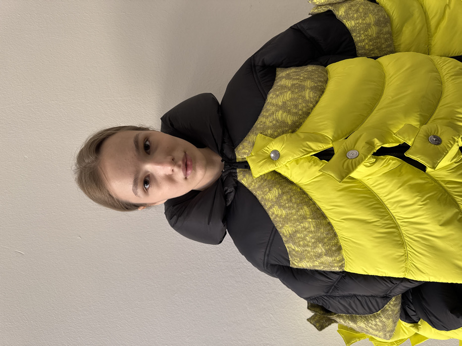 A young adult female with light skin and light brown hair pulled back stands against a plain, light-colored wall in what is identified as an indoor setting in Milan, Italy. She faces forward, making direct eye contact with the camera, displaying a neutral facial expression. The subject is wearing a prominent, multi-textured puffer jacket. The garment features a black upper section with a high, padded collar. The central and lower portions of the jacket are bright yellow, constructed from large, layered, puffed panels secured by visible silver-colored snap buttons. Integrated within the design are sections of a textured, muted yellow-green fabric displaying a repetitive, intricate pattern, visible on the chest and extending to the shoulders and sides. A black zipper is visible on the front closure. The lighting is even, indicating controlled illumination.