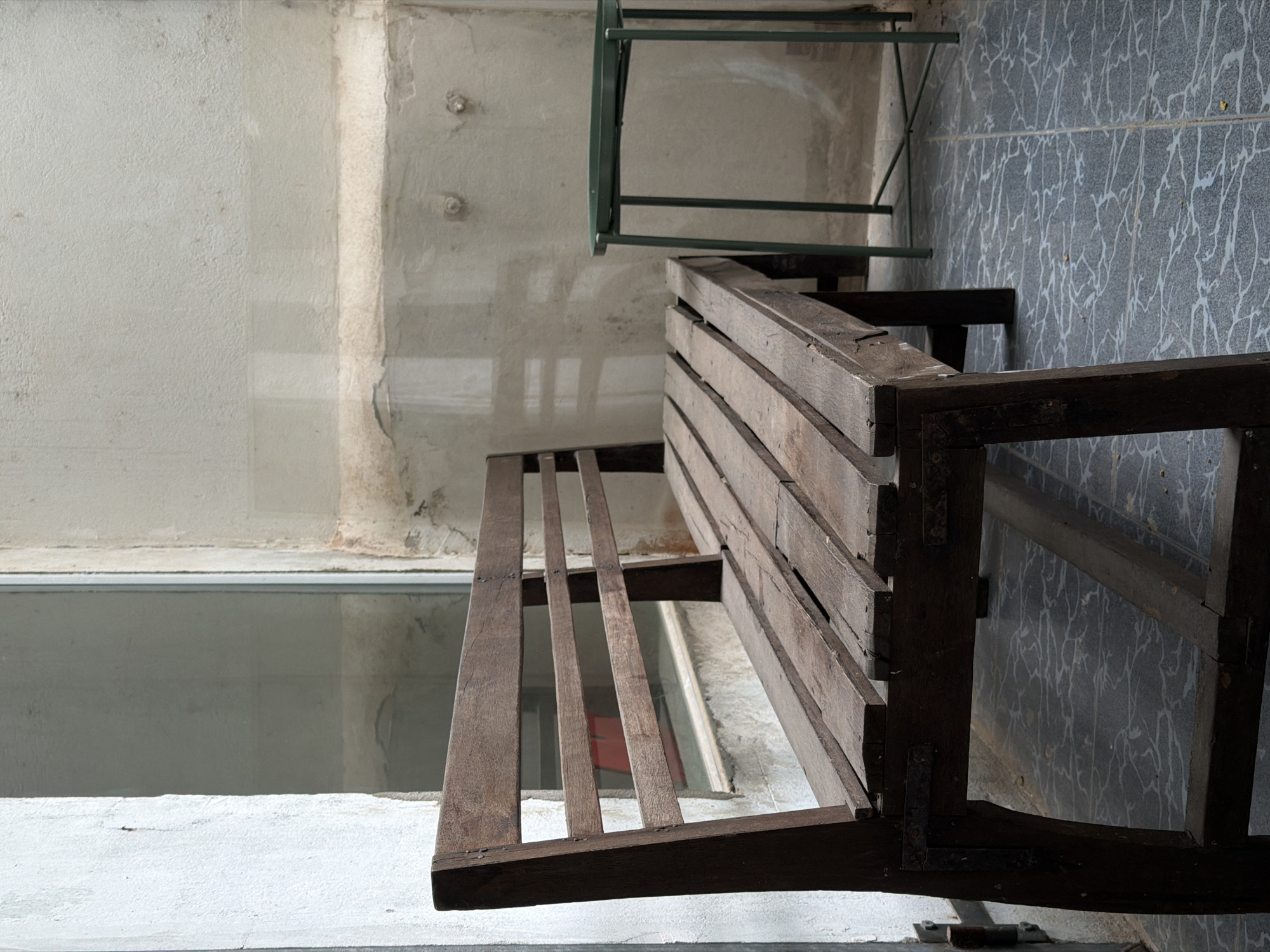 An outdoor, covered seating area in Da Nang, Vietnam, features a rustic wooden bench and a small green side table. The bench, made of weathered, dark wood planks, is positioned prominently in the foreground, facing slightly to the right. Its backrest consists of horizontal wooden slats. To the right of the bench, a slender, dark green metal side table with a tray-like top stands. The flooring is tiled with a dark gray pattern featuring white, irregular lines. In the background, a wall with a textured, light-colored finish is visible, marked by some water stains and what appear to be two small, round fixtures. A large window or glass partition separates this area from another space, reflecting the light and some indistinct shapes. The overall impression is one of a simple, slightly aged outdoor space.