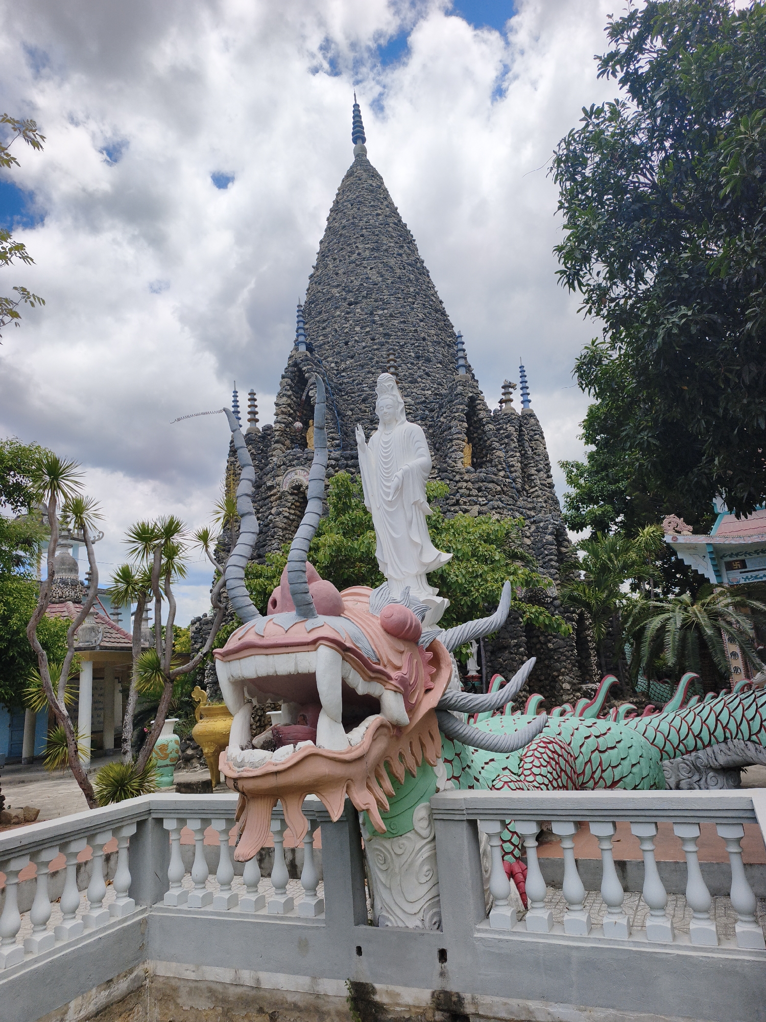 This image captures a view of the Dragon Pagoda (Chùa Long Sơn) in Cam Ranh, Vietnam. In the foreground, a colorful, ornate dragon sculpture is prominently displayed. The dragon's head, with its mouth open, is pink and white, with large tusks. Behind the dragon's head, a white statue of a serene female figure stands on a pedestal. The dragon's body, visible further to the right, is green with red scale detailing.
A white balustrade with pink accents runs across the lower portion of the image, separating the foreground elements from a sunken, possibly decorative, area. Beyond the dragon and statue, a large, tiered pagoda constructed from dark stones and resembling a mountain peak dominates the background. Smaller pagodas with blue decorative spires are visible on the sides of the main structure. Lush green trees and palm trees surround the pagoda. The sky above is partly cloudy, with patches of blue sky peeking through. In the mid-ground, to the left, there are other temple structures with light-colored walls and roofs. The overall scene depicts a religious and cultural site with intricate artistic elements.