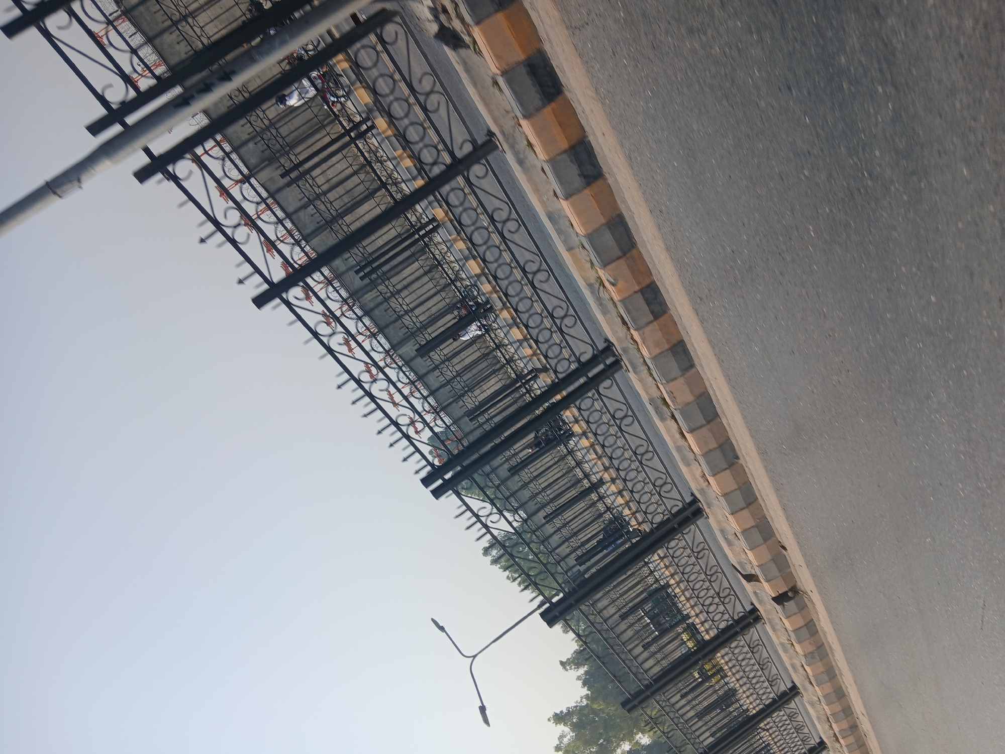 A low-angle, tilted shot captures a street scene in Peshawar City Tehsil, Pakistan. In the foreground, an asphalt road occupies the bottom half of the frame, with a curb lined with alternating yellow and grey concrete blocks. Beyond the curb, a black metal fence with decorative scrollwork extends diagonally across the image, creating a barrier. This fence appears to be enclosing a walkway or elevated area. Through the fence, several parked bicycles, mostly orange and red, are visible. A street light with two lamps stands on the left side of the frame, near the fence. In the background, a clear, pale sky dominates the upper portion of the image, suggesting daytime. The overall scene depicts an urban environment with elements of street infrastructure and transportation.