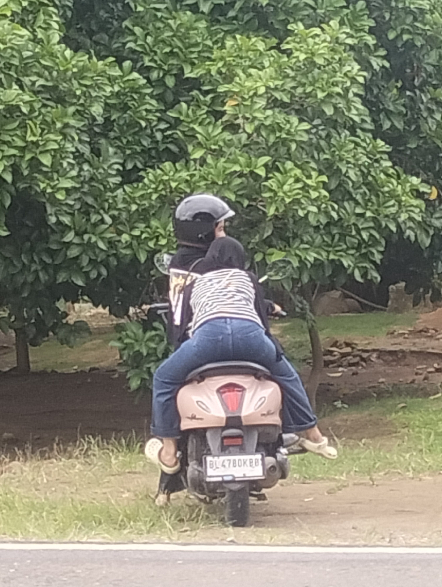 Two individuals are riding a light pink scooter on a paved road in a rural or suburban area, possibly in Teupinreusep, Indonesia. The person in the back, wearing a black and white striped shirt and blue jeans, is seated facing forward. The person in the front, wearing a black jacket and a black helmet, is also seated facing forward. The scooter has a license plate with the characters "BL 4780 KBB." The background consists of lush green trees and foliage, suggesting an outdoor environment. The ground is a mixture of dirt and grass.