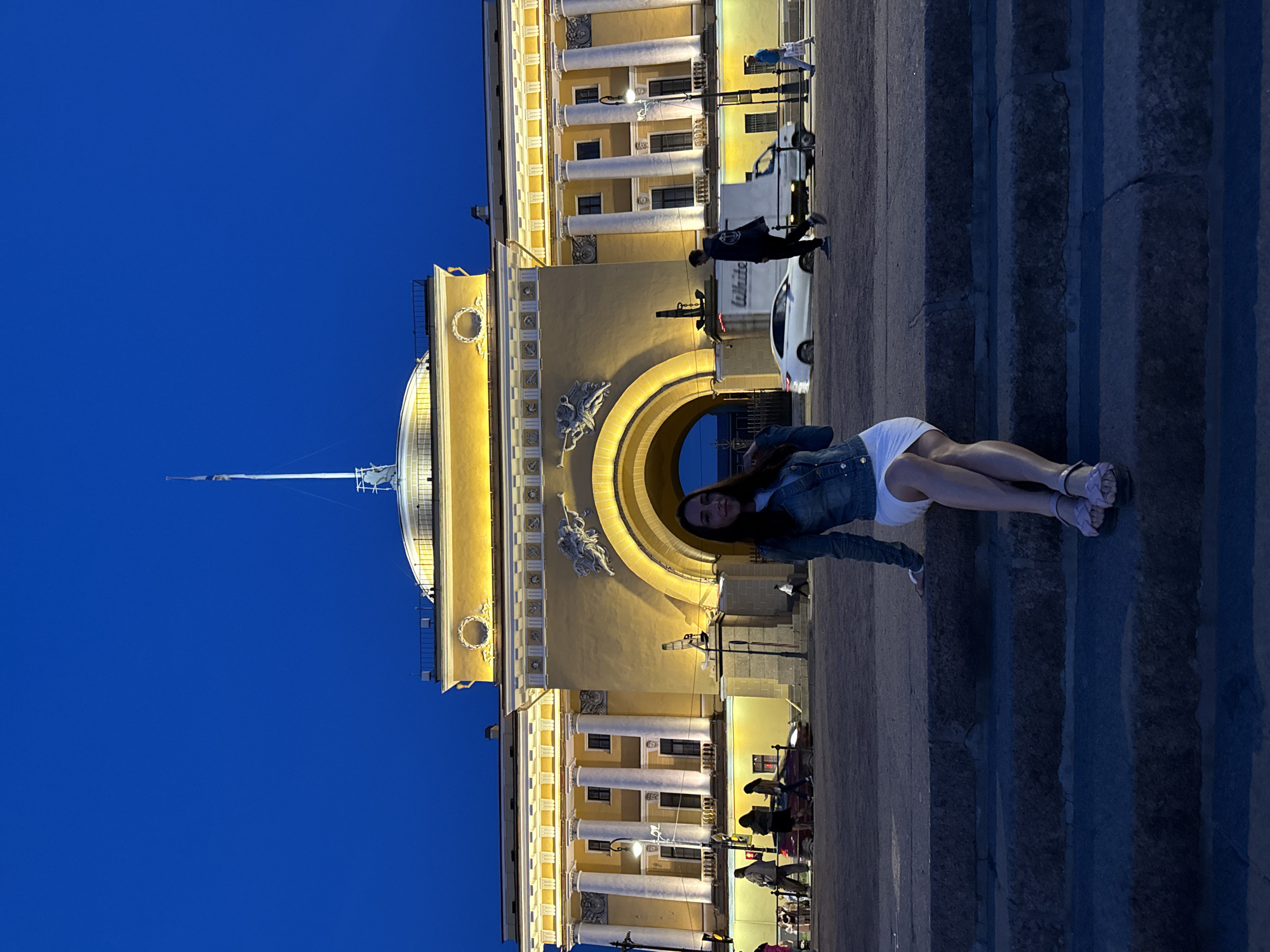 This photograph captures a woman posing in front of the illuminated Admiralty Building in Saint Petersburg, Russia, during twilight or the "blue hour." The sky is a deep, clear blue, providing a stark contrast to the warm, golden glow of the building's facade, which is brightly lit by artificial lights.
The main subject, a woman, stands on the right side of the frame, facing slightly left and smiling towards the camera. She wears a light blue denim jacket over a white top or short dress, paired with light-colored, possibly lavender or gray, strappy sandals. Her long dark hair is down. She is positioned on a dark, paved surface, possibly steps, which recedes into the foreground.
Behind the woman is the grand, Neoclassical architecture of the Admiralty Building. Its central feature is a massive arched entryway, flanked by colossal white columns and adorned with intricate sculptural reliefs above the arch. To the left, a prominent golden dome with a slender, vertical spire rises high into the sky. The building's facade is detailed with multiple rows of white columns, decorative friezes, and numerous windows.
In the background, visible through and around the central arch, are several vehicles, including a white delivery van with the partial text "White..." visible on its side, and a white car. Other indistinct figures of people can be seen further in the distance, indicating a public urban setting. The overall scene conveys a serene evening atmosphere, emphasizing the architectural splendor of the landmark.