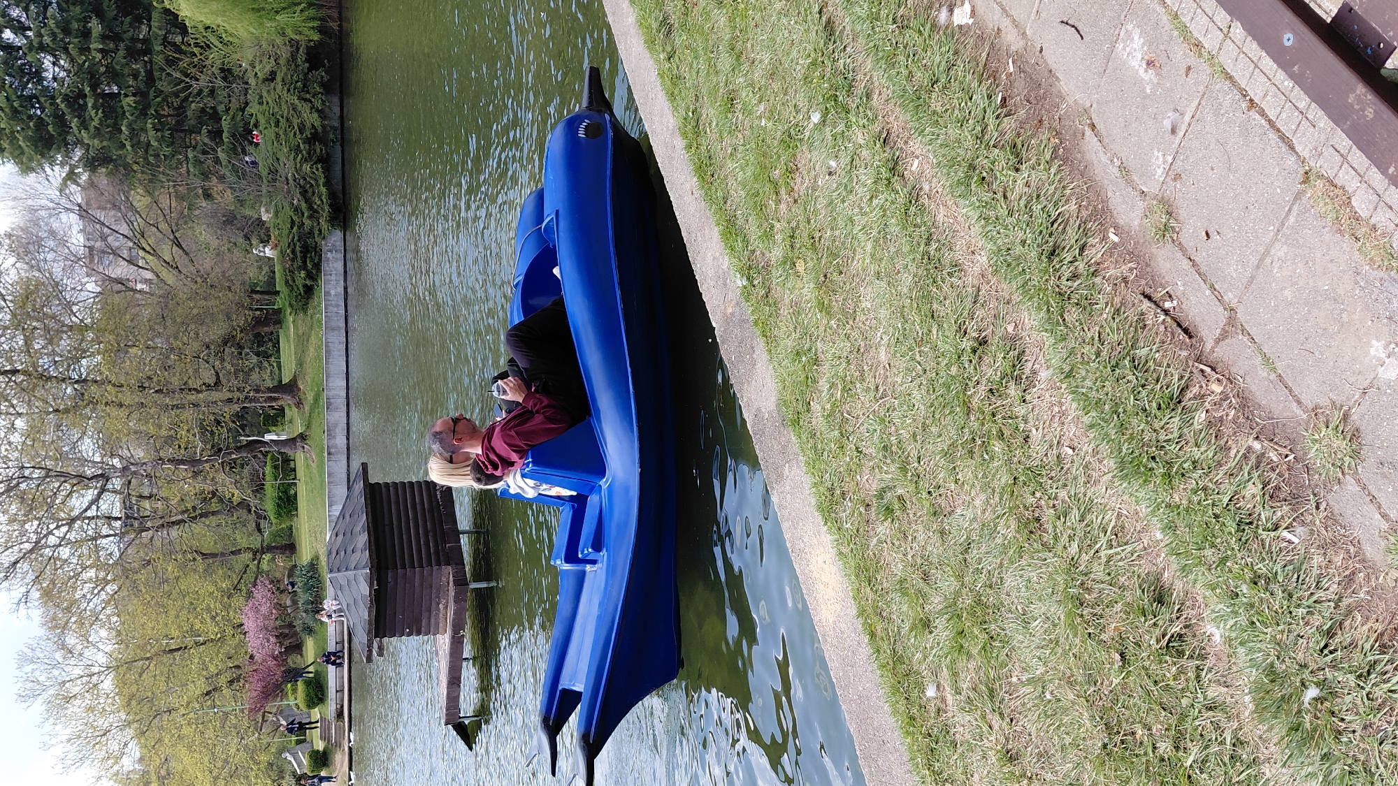 The image captures a tranquil scene by a body of water, possibly a pond or a small lake, in an urban park. A blue pedal boat is docked at the water's edge. Inside the boat sits a person wearing a maroon top and dark pants, holding a camera and looking towards the water. A concrete bank lined with grass is adjacent to the boat.
On the other side of the water, the scene opens up to reveal a small wooden boathouse, trees with sparse foliage, and other people strolling around the park. The location seems to be an urban park due to the presence of the managed landscape, public seating, and recreational facilities. The city is Unknown, Unknown.