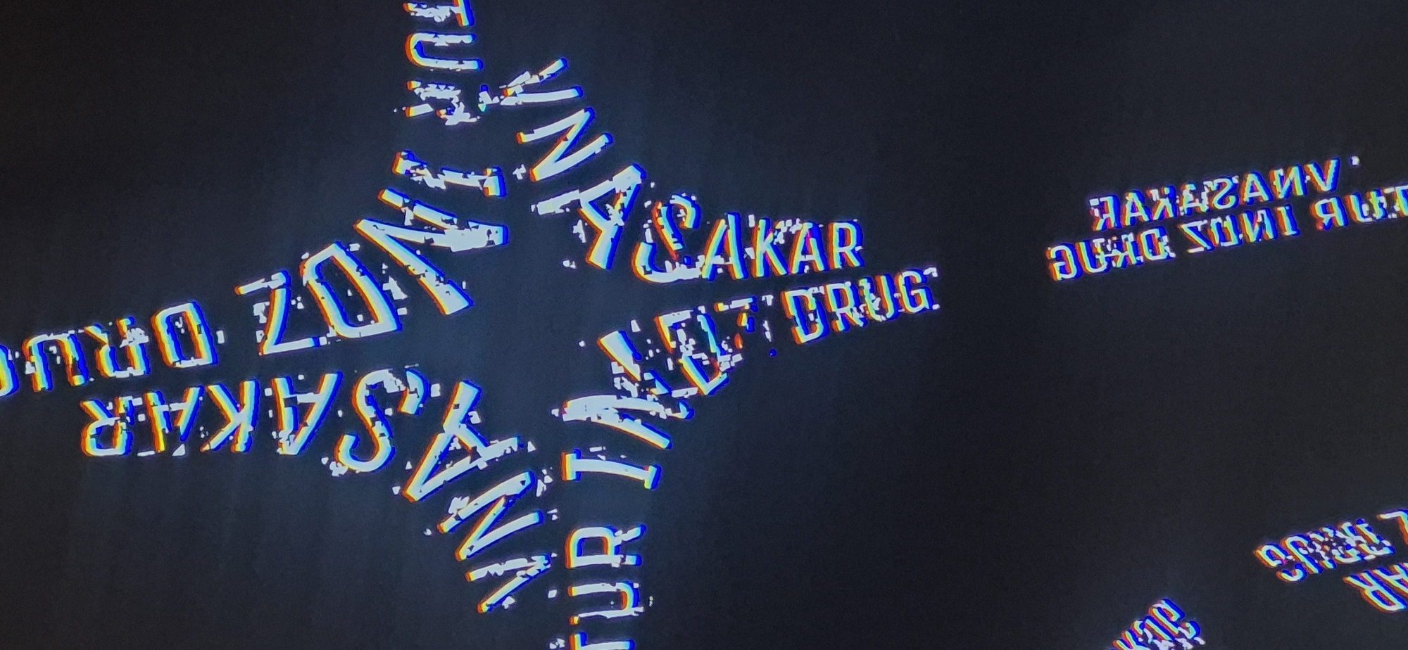 The image features text with a distorted, glitching effect, set against a dark background. The text includes the words "VANASAKAR", "SAKAR", "DRUG", and other fragments, arranged in an overlapping, radiating pattern. The location is not discernible from the image. No people or specific objects are visible.