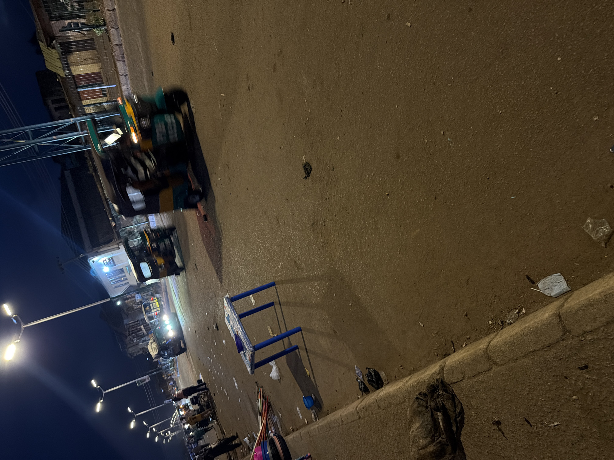 This image captures a street scene at night in Bukuru, Nigeria. The road appears wet, suggesting recent rain or a damp environment. Several streetlights illuminate the area, casting long shadows. A blue plastic table with a blue bowl on top sits in the middle of the road, surrounded by scattered litter, including plastic bags and what appear to be discarded food wrappers. To the left, a concrete sidewalk is visible, with more debris, including what looks like matted hair or fur, and a crumpled plastic bag near the curb.
In the background, two auto-rickshaws, commonly known as "keke napep" in Nigeria, are driving on the road. One is in the foreground, with its lights on and motion blur indicating movement. The other is further back, also illuminated. People are visible on the left side of the road, seemingly gathered near stalls or shops. Buildings line the street, some with illuminated windows. The overall impression is of a bustling urban environment at night, with evidence of commercial activity and the common presence of street litter.