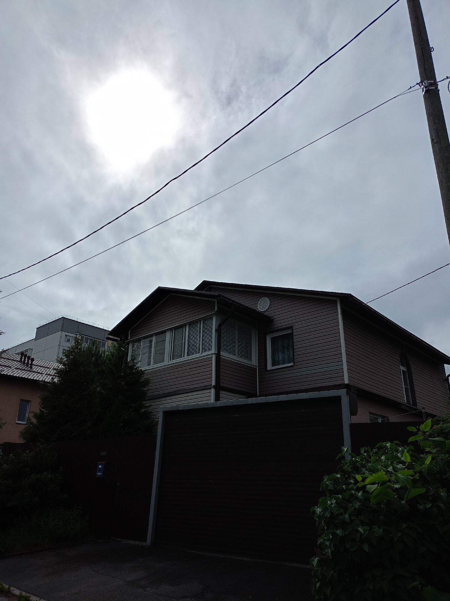 An overcast day is captured in this outdoor scene, with the sun partially obscured by clouds, creating a bright, diffused light source in the upper left quadrant. The image focuses on the exterior of a two-story building, likely a house, with brown siding and white trim. A large, dark garage door dominates the lower portion of the frame. Adjacent to the house, a dense evergreen tree is visible. In the background, another building with a grey facade and multiple windows can be seen. Power lines stretch across the sky, supported by a utility pole on the right side of the image. The ground in front of the garage is paved.