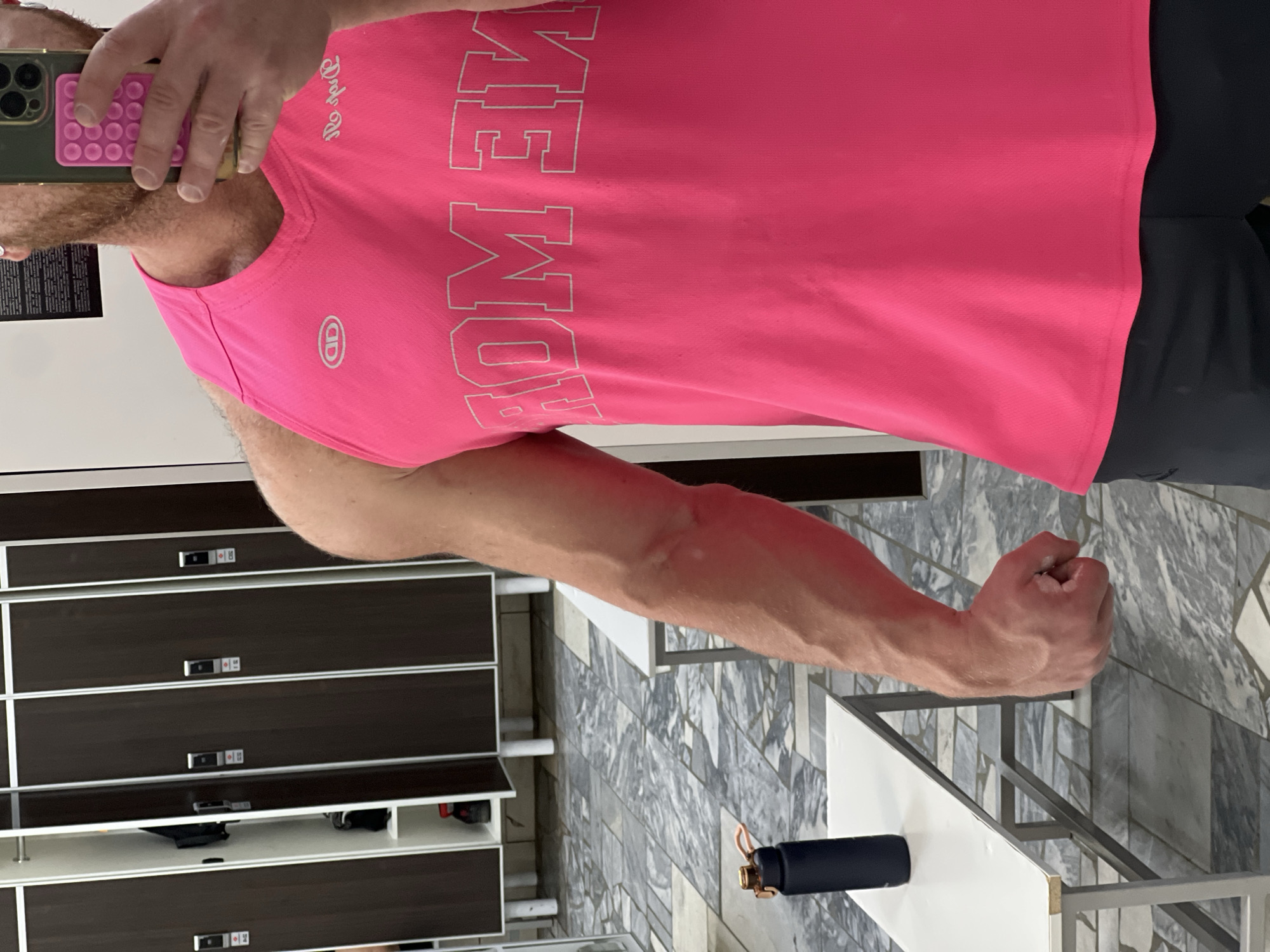 The image depicts a male individual taking a mirror selfie in a locker room located in Chaikovsky, Russia. The individual is visible from the mid-chest to the upper thighs. They are wearing a bright pink sleeveless top with white lettering, which appears reversed in the reflection, showing "MOM TIF" and "ME MOM." A circular logo is present on the upper left chest. Dark shorts are visible below the top.
The individual's right arm is flexed, displaying pronounced musculature and visible veins, with the hand clenched into a fist. A green-cased smartphone, equipped with a pink circular pop-socket and multiple camera lenses, is held in the individual's left hand, used to capture the image in the mirror. Partial visibility of a reddish beard is present on the reflected face.
The background shows a row of dark brown lockers with white vertical frames and silver-colored locks or handles. Several locker doors are slightly ajar, revealing white interior shelving and some dark items. One locker on the far left is fully open. Two benches with white tops and light grey metal frames are positioned in the foreground and midground. A dark blue or black water bottle, featuring a copper-colored ring and a light-colored strap, rests on the bench in the immediate foreground. The floor is covered with patterned tiles in shades of grey, white, and black. A white wall is visible behind the lockers, with a dark rectangular object mounted higher on the wall to the right.
