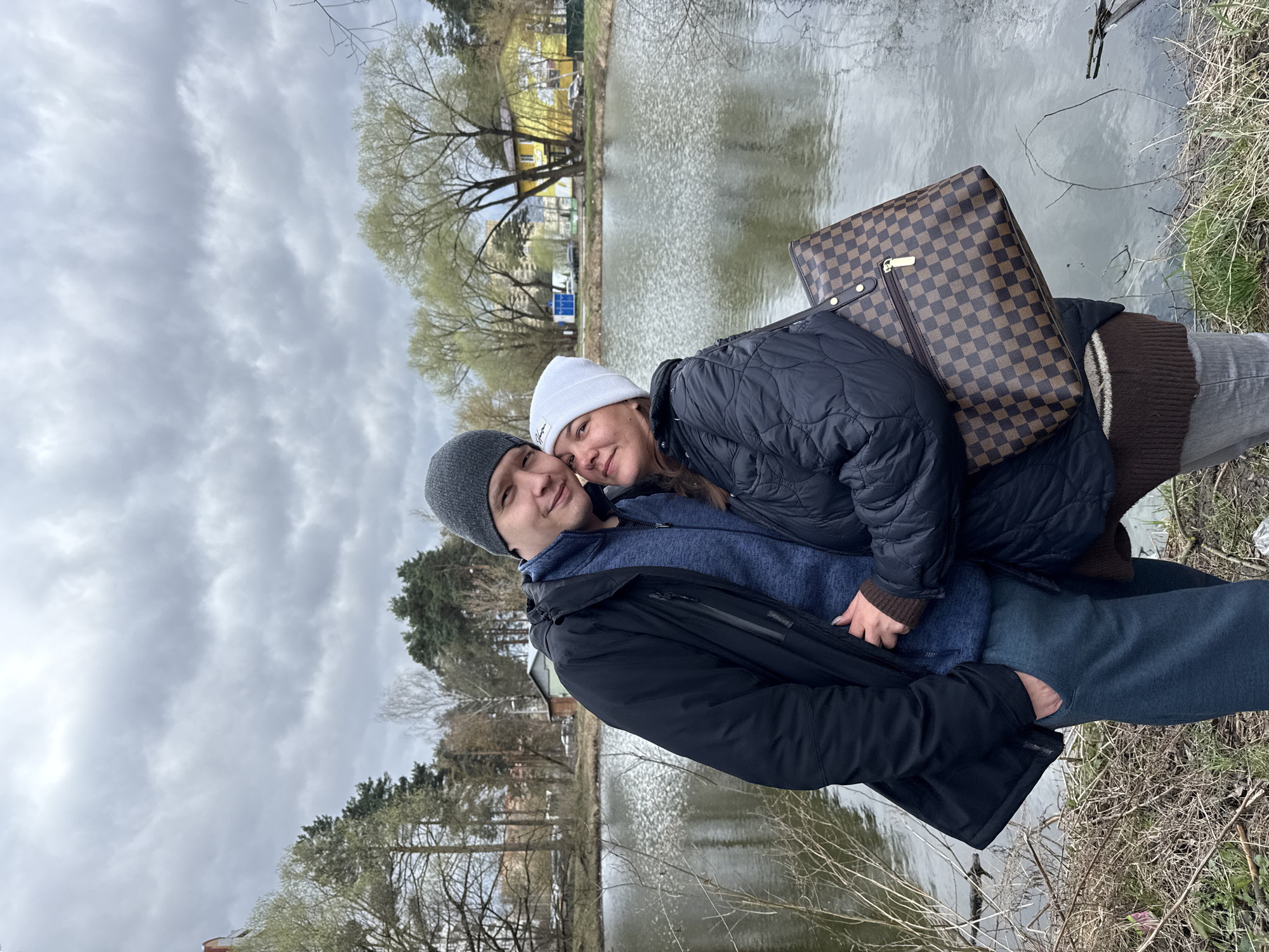 Two individuals are posing for a photograph outdoors, likely near a body of water. The man on the left wears a gray beanie, a blue sweater, and a dark jacket. The woman to his right is wearing a white beanie and a quilted black jacket. A checkered brown handbag rests on her lap. The setting includes a pond or lake, with trees and buildings visible on the opposite shore. The image's origin suggests the location may be in Dmitrov, Russia. The sky is overcast. The couple appears to be standing or leaning against something near the edge of the water.