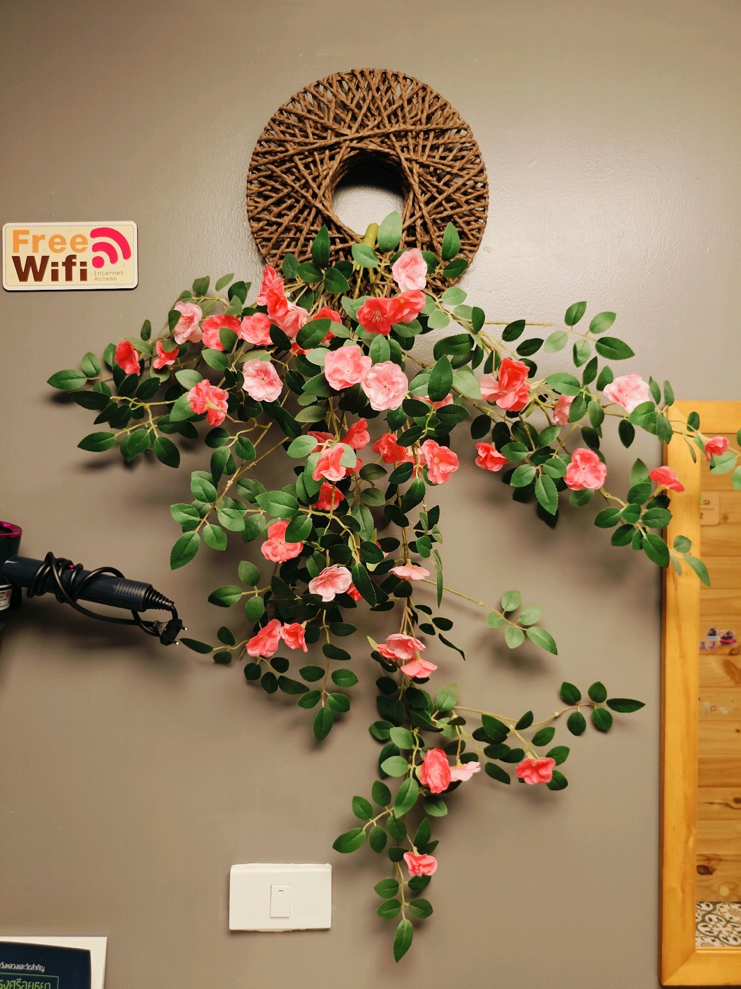 The image depicts a wall decorated with a woven wreath and artificial pink flowers. To the left of the wreath is a "Free Wifi" sign. A hair dryer is visible on the lower left side of the image, plugged in and resting against the wall. A white light switch is present on the lower middle section of the wall. On the right side of the image, a wooden frame with some items visible inside is present. The overall setting appears to be indoors, possibly in a salon or a guesthouse in Phra Nakhon Si Ayutthaya City Municipality, Thailand, given the presence of the hair dryer and the wifi sign.