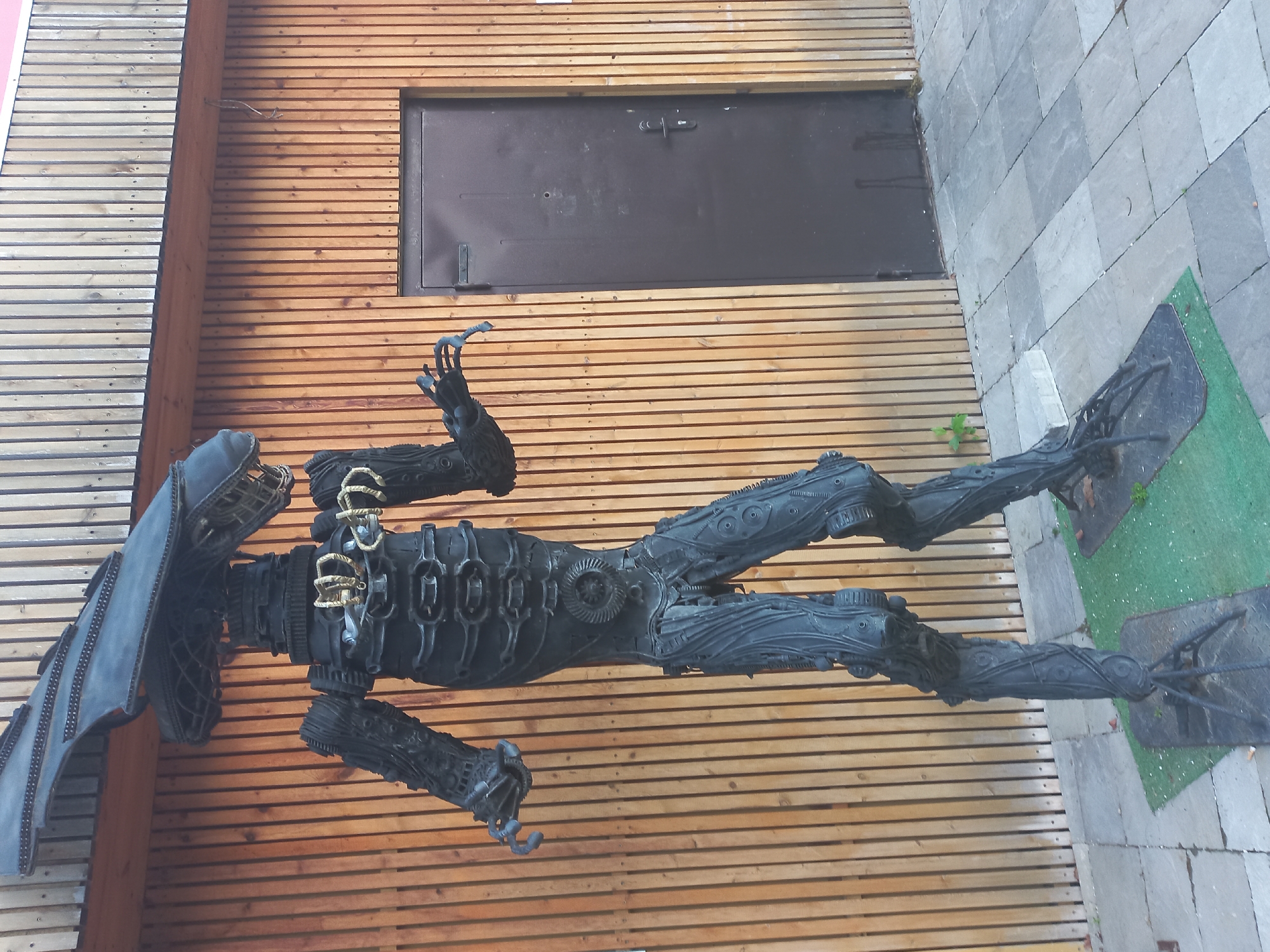 The image depicts a sculpture of a Xenomorph from the "Alien" film franchise, located in Sochi, Russia. The sculpture is black and appears to be made of recycled metal parts. It is positioned in front of a modern building with horizontal wooden planks for siding and a black metal door. The sculpture is lying horizontally, with its head on the left and feet on the right. Its limbs are articulated and the body is detailed with mechanical and organic elements. The base of the sculpture rests on a combination of paving stones and artificial turf. The surrounding environment suggests an urban setting.
