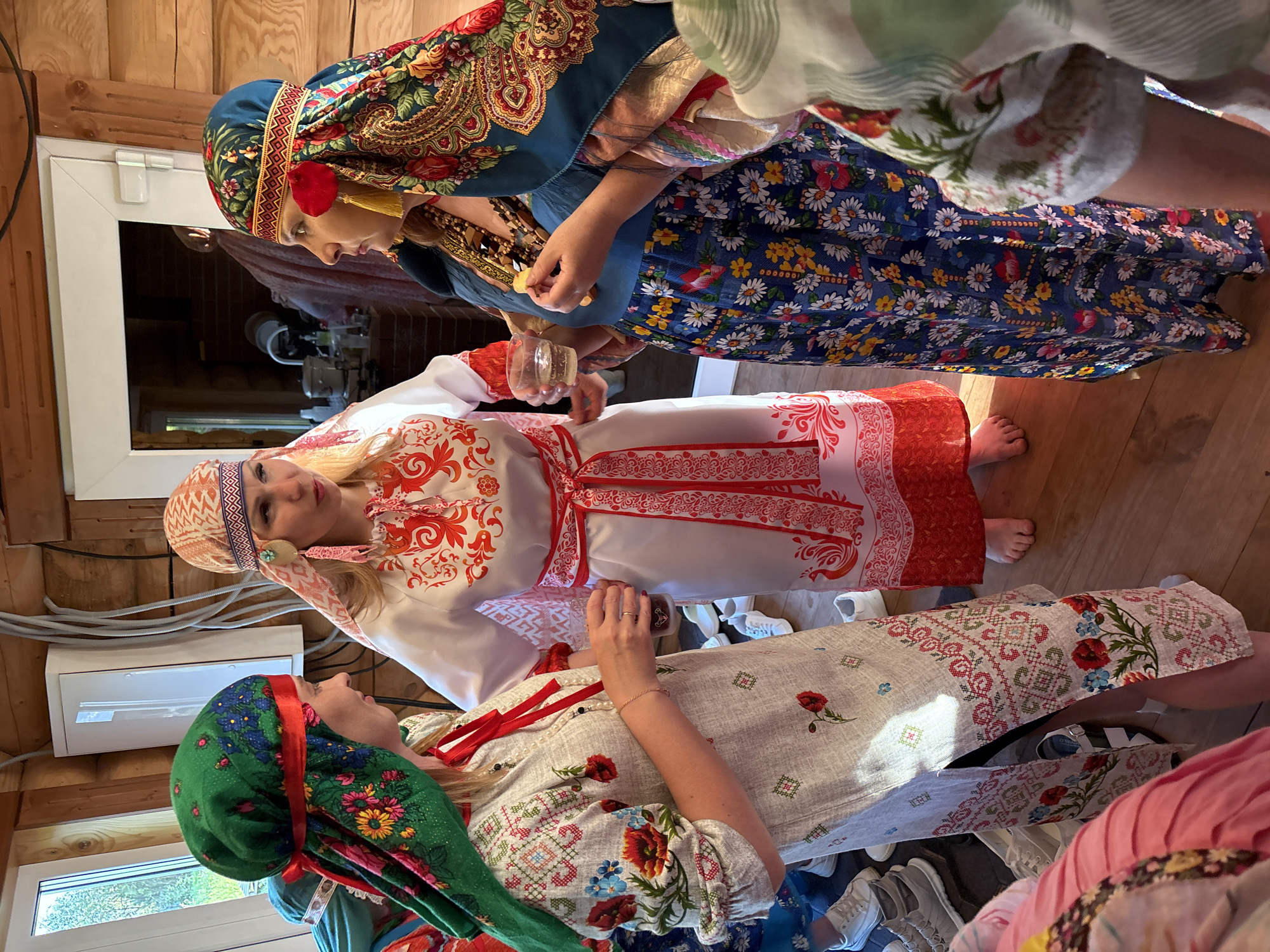 Three women are standing in a room with wood-paneled walls, wearing traditional Russian clothing.
The woman on the top wears a blue dress with a floral pattern and a matching headscarf. She's holding food and a glass. The woman in the middle wears a white dress with red ornamental patterns, and a matching headscarf. The third woman, at the bottom, wears a white dress with a floral pattern and a green headscarf. All the women are interacting with each other.
The room has windows and an electrical panel on the wall. There is clutter on the floor, including shoes.
The location is Markovo, Russia.