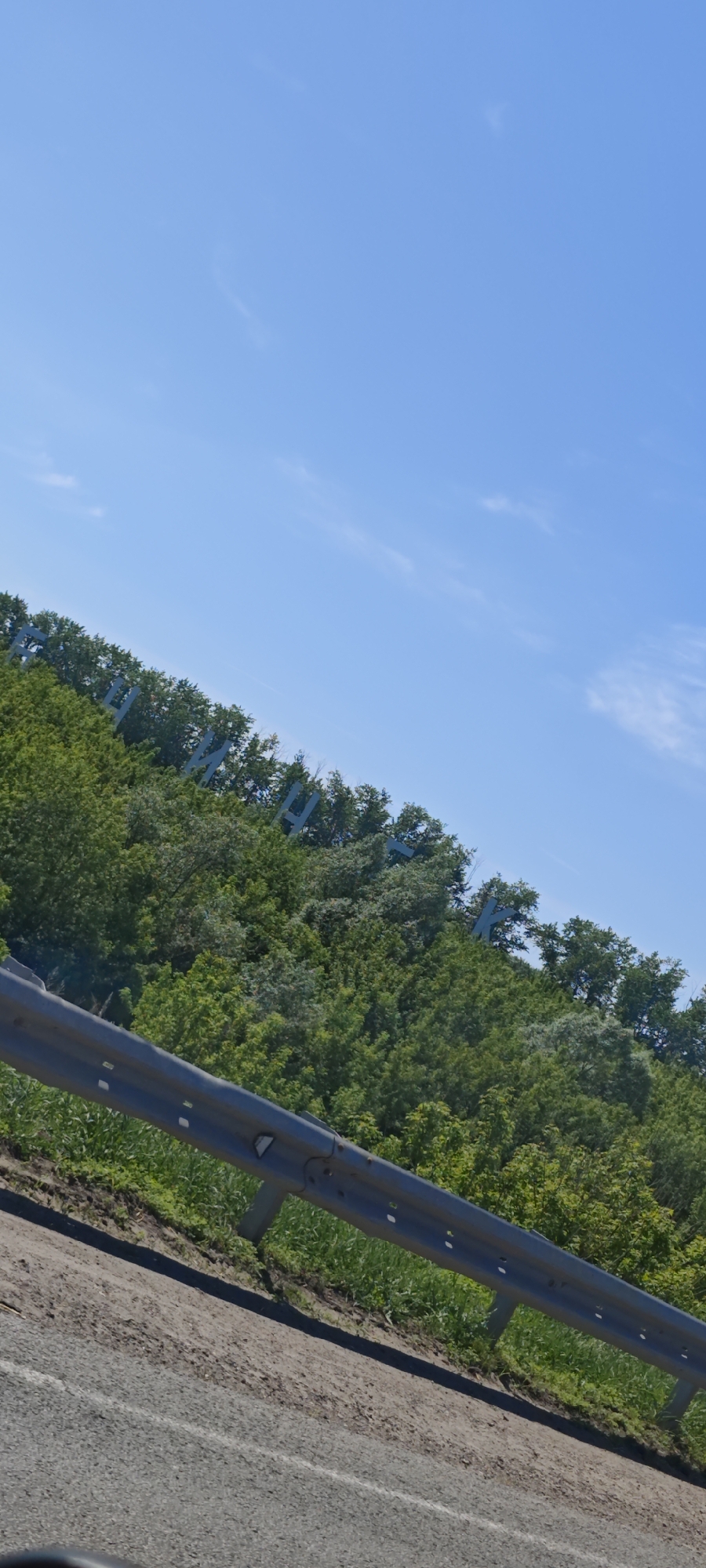 The image shows the Buinsk sign on a hill, viewed from what appears to be a roadway. A guardrail is visible in the foreground. The sky is blue with some clouds. The hillside behind the road is covered in green trees, through which the letters of the city name, "БУИНСК" (BUINSK), are positioned.