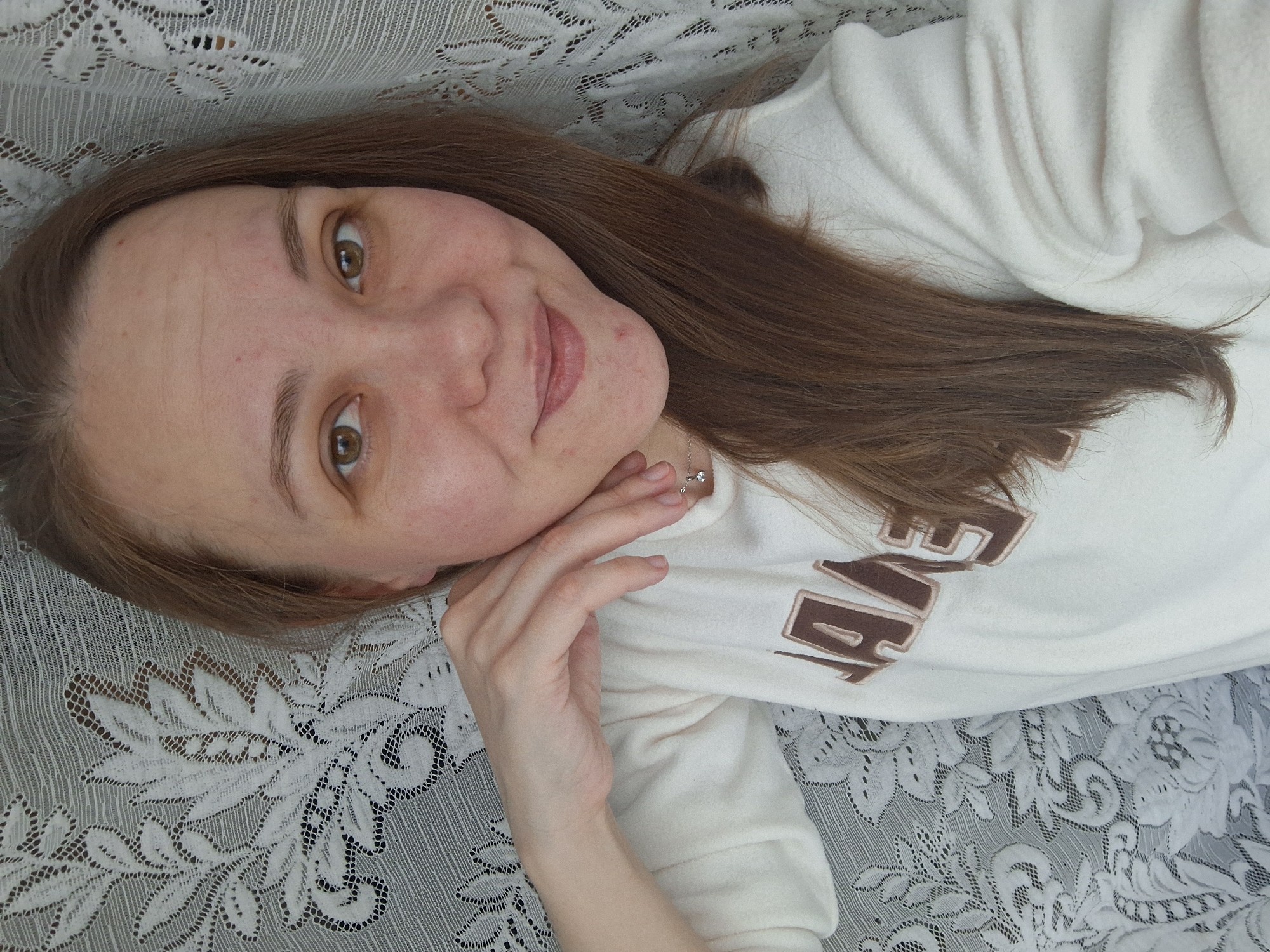 A fair-skinned woman with brown hair is lying on a white, lace-covered surface. She is wearing a white sweatshirt with brown lettering. She has brown eyes and is smiling slightly. Her left hand is near her face. The location is Chaikovsky, Russia.