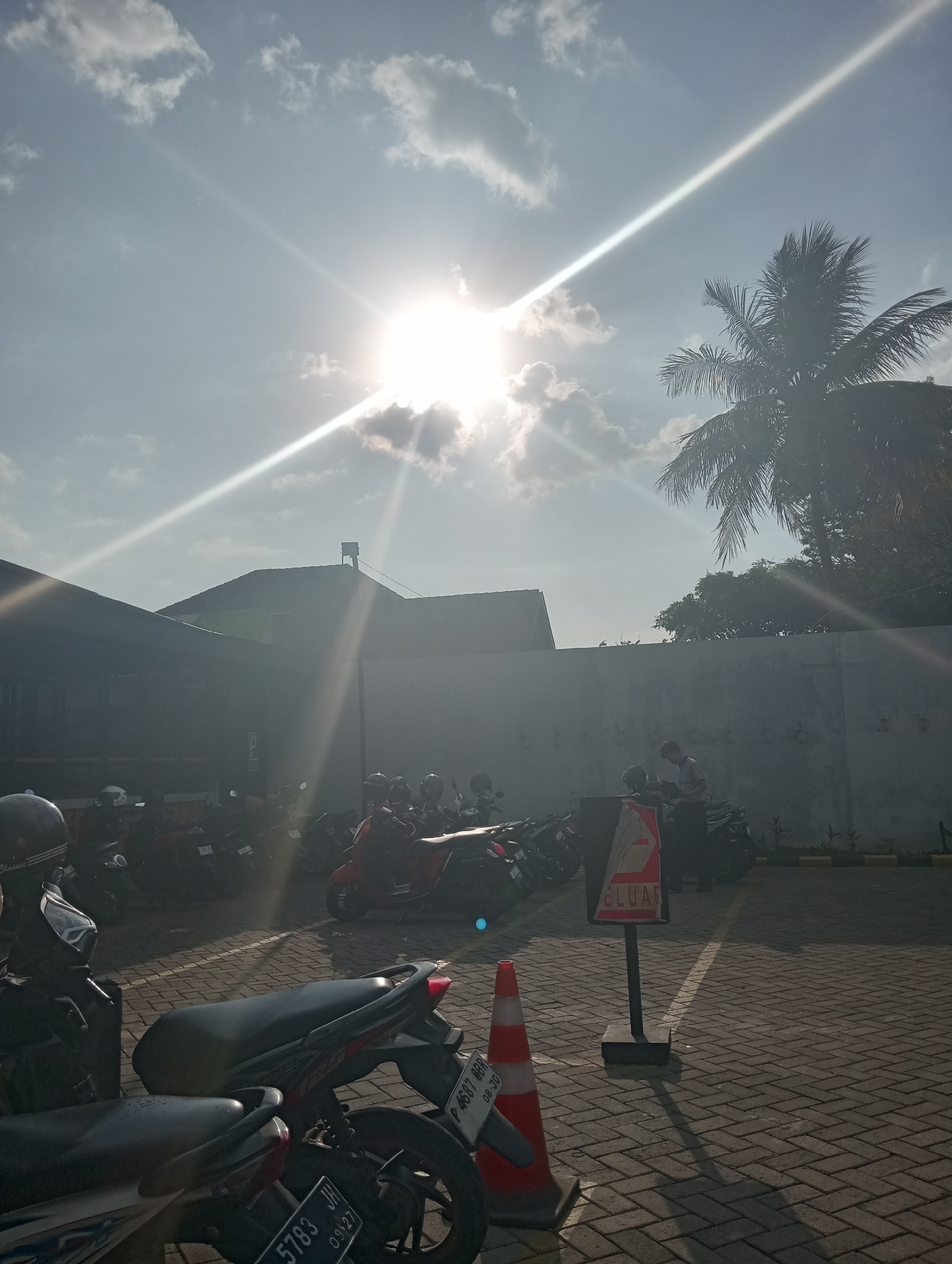 This is a photograph taken on a sunny day, likely in Jember, Indonesia, given the presence of motorcycles and the context of the provided information. The image depicts an outdoor parking area paved with interlocking bricks. The sun is prominently featured in the sky, casting strong lens flares and light rays. A silhouette of a palm tree is visible on the right side of the frame.
In the foreground, several motorcycles are parked. One motorcycle in the immediate foreground is black and grey, with visible license plates. To its right, another motorcycle is partially visible. Further back, a row of parked motorcycles stretches into the middle ground.
A light grey wall runs horizontally across the middle of the image, separating the parking area from a building or structure behind it. A sign with the word "KELUAR" (which means "EXIT" in Indonesian) is mounted on a post in the parking area. An orange traffic cone is also present near the sign.
In the background, a low-rise building with a dark roof is visible, partially obscured by the bright sunlight. The sky is bright blue with scattered clouds. The overall scene suggests a typical day in an urban or semi-urban environment in Jember, Indonesia, focusing on a motorcycle parking lot under strong sunlight.