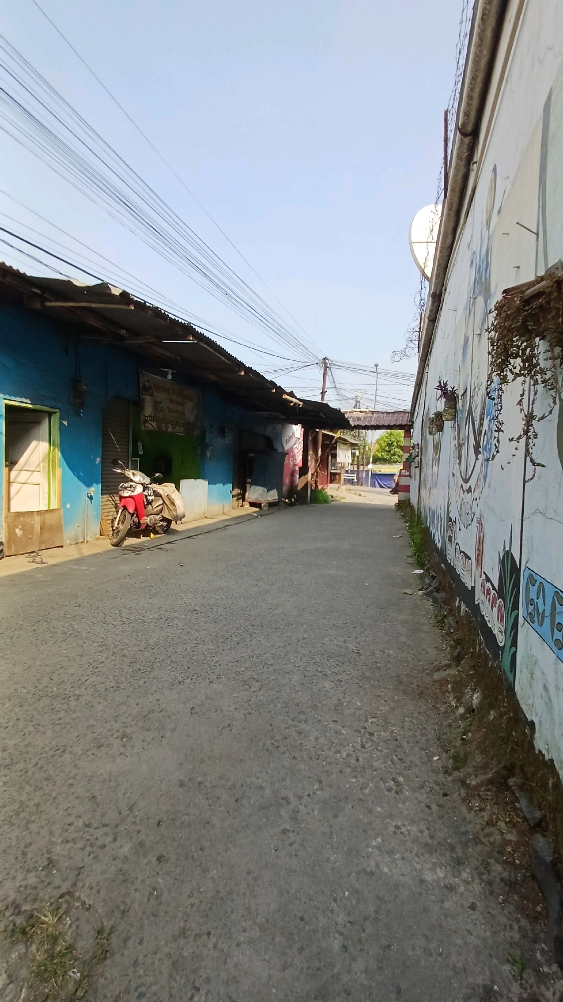 The image depicts a street scene in Gunung Putri, Indonesia, characterized by a narrow asphalt road flanked by buildings on the left and a mural-decorated wall on the right. The buildings on the left appear to be shops or residences with blue and green facades, some with corrugated metal roofs. A red and white motorcycle with a large sack on its back is parked in front of one of the blue buildings. Overhead, a dense network of electrical wires crisscrosses the sky. The wall on the right features a colorful mural with various figures and designs, and a satellite dish is mounted on it. In the background, the road continues, passing under an arched structure, with glimpses of greenery and more buildings beyond. The lighting suggests it is daytime, with clear skies. The overall impression is of a modest urban or semi-urban environment.