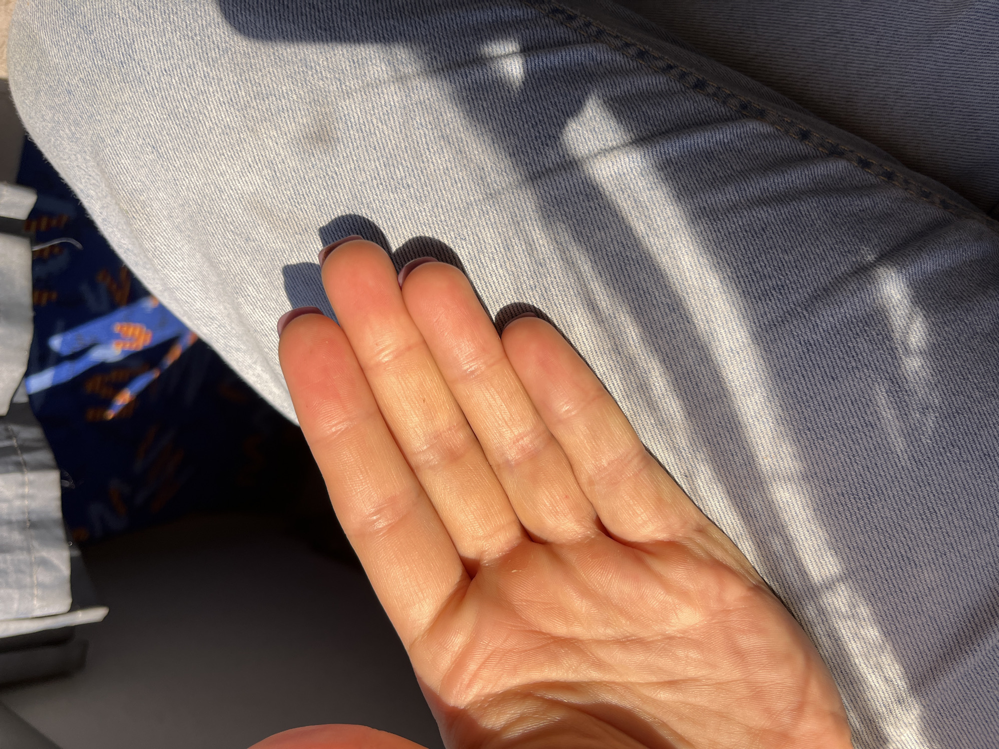 A close-up shot focuses on a person's left hand, palm up, resting on light blue denim jeans. The hand appears to be illuminated by warm, angled sunlight, creating shadows that highlight the texture of the skin. The fingernails are painted a dark color. The background is blurred, showing a dark area and a hint of fabric with a pattern of blue and orange. The scene is not identifiable as being in Myadzyel, Belarus.