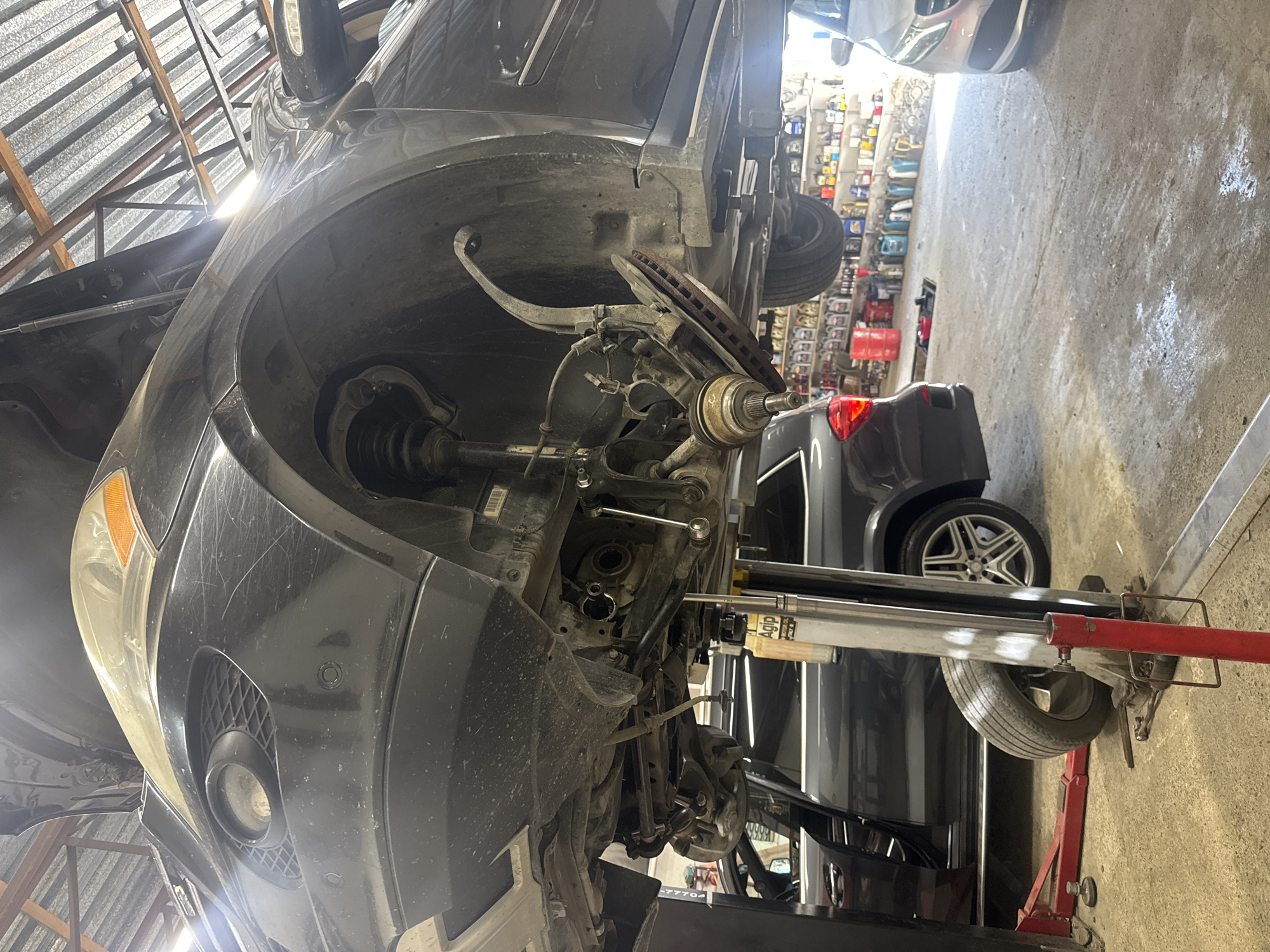An undercarriage view of a black Mercedes-Benz SUV is prominently featured, hoisted on a hydraulic lift in a workshop setting. The front wheel assembly is partially disassembled, with the brake rotor exposed. In the background, another dark-colored Mercedes-Benz sedan with its door open is parked. Shelves stocked with various automotive fluids and products line the back wall. The scene appears to be a car repair shop. No specific location in Baku, Azerbaijan is visible, nor are any people depicted.
