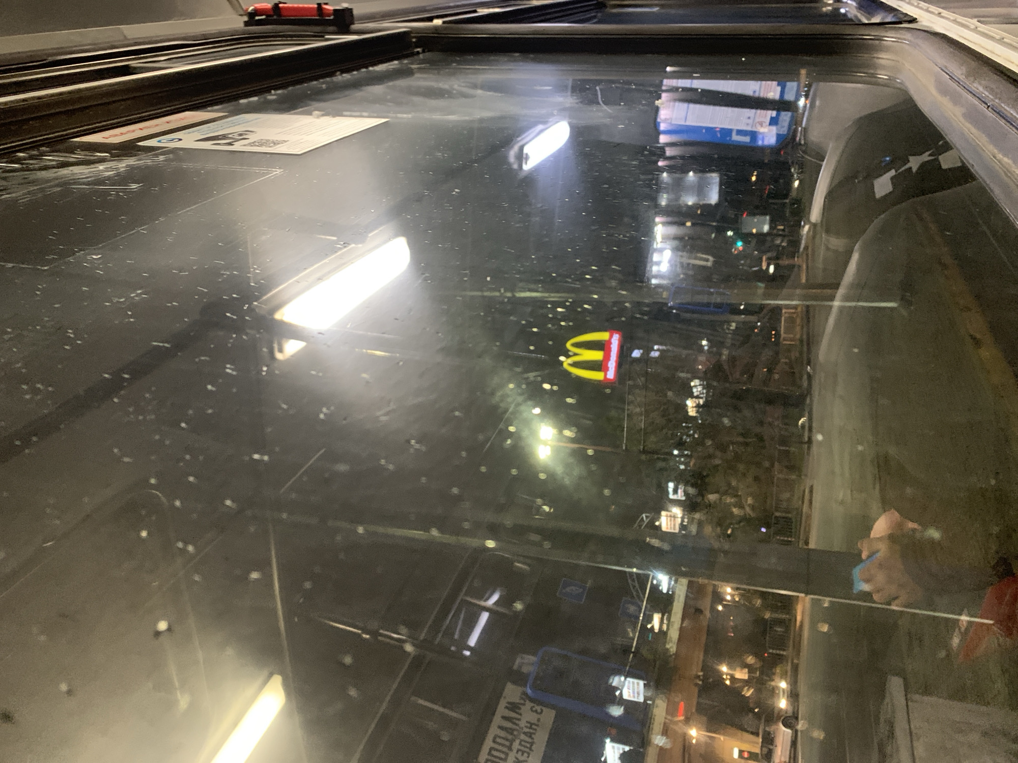 The image shows a nighttime view of Sofia, Bulgaria, as seen through a rain-streaked glass window, likely from inside a building or vehicle.
Outside the window, the city is illuminated by streetlights and signs. A McDonald's logo is visible among the lights. There are traffic signs written in Cyrillic. Other signs with Cyrillic text are present.
Inside, reflections of interior lights and objects are visible on the glass surface. There is a card on the left side of the image.
