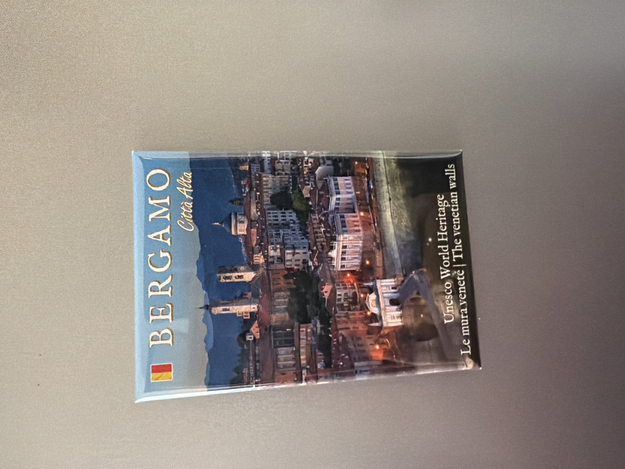 A rectangular magnet displays a nighttime image of the city of Bergamo, Italy, with text indicating it is a UNESCO World Heritage site. The magnet is affixed to a light-colored, textured surface, likely a refrigerator. The top of the magnet features the word "BERGAMO" in large, serif font, with the Italian phrase "Città Alta" beneath it. The image itself is a cityscape with illuminated buildings, spires, and a domed structure, set against a dark blue sky and silhouetted mountains. The bottom of the magnet has text that reads "Unesco World Heritage Le mura venete | The venetian walls".