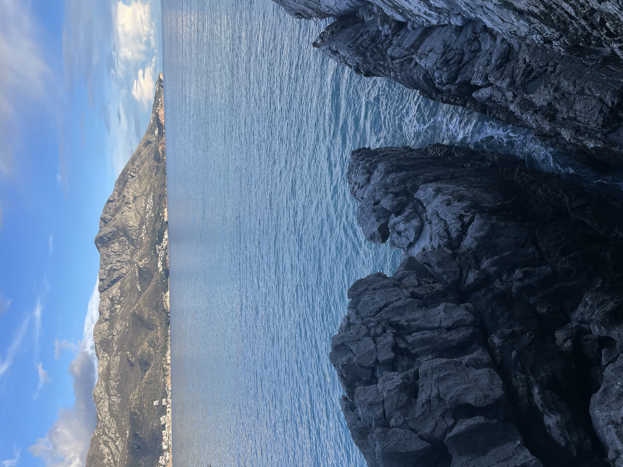 The image depicts a panoramic coastal landscape in Budva, Montenegro. In the immediate foreground, dark, rugged rock formations with fractured surfaces dominate the lower portion of the frame. Between two prominent rock masses on the right, a narrow channel of blue-green water exhibits minor wave action. The midground consists of a wide expanse of calm, blue-green sea water, showing subtle ripples across its surface. In the background, a chain of mountains, appearing rocky and sparsely vegetated, stretches horizontally. Along the base of these mountains, on the distant coastline, scattered white and light-colored buildings indicate a human settlement. The sky above is partially clear with patches of blue and scattered white and gray cumulus clouds. No people, animals, or specific actions are present in the scene.