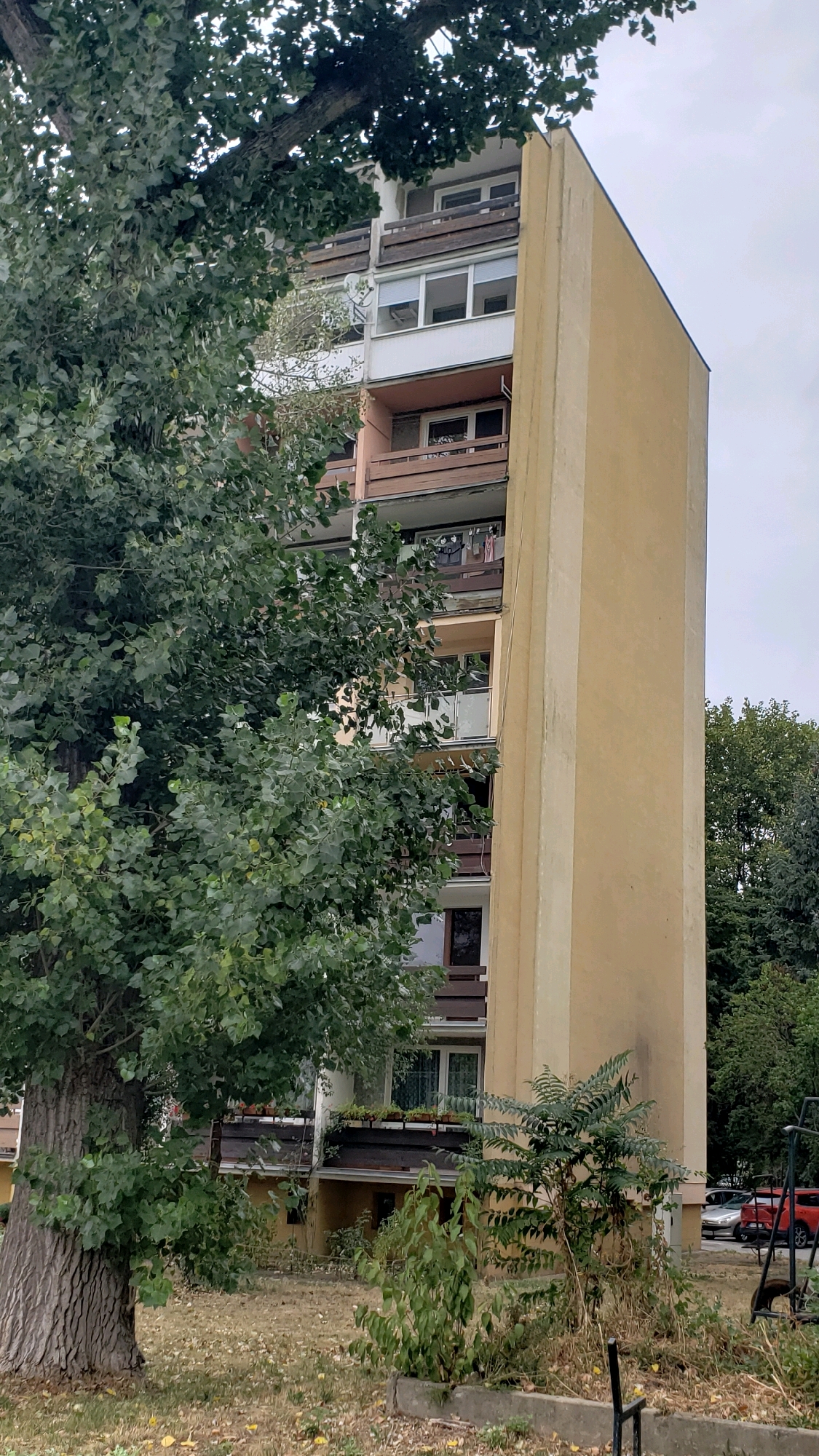 This is a full-frame, eye-level, outdoor, daylight photograph of a tall, yellow apartment building in the city of Prešov, Slovakia. A large tree with dark green leaves stands in the foreground to the left, partially obscuring the building's facade. The building features multiple balconies with wooden railings, and several windows are visible, some with closed blinds. There are also some potted plants on the balconies. The ground in front of the building is covered with dry grass and fallen leaves. A few vehicles are parked in the background, and some smaller trees and bushes are present. The sky is overcast. The overall impression is one of a residential area on a cloudy day.