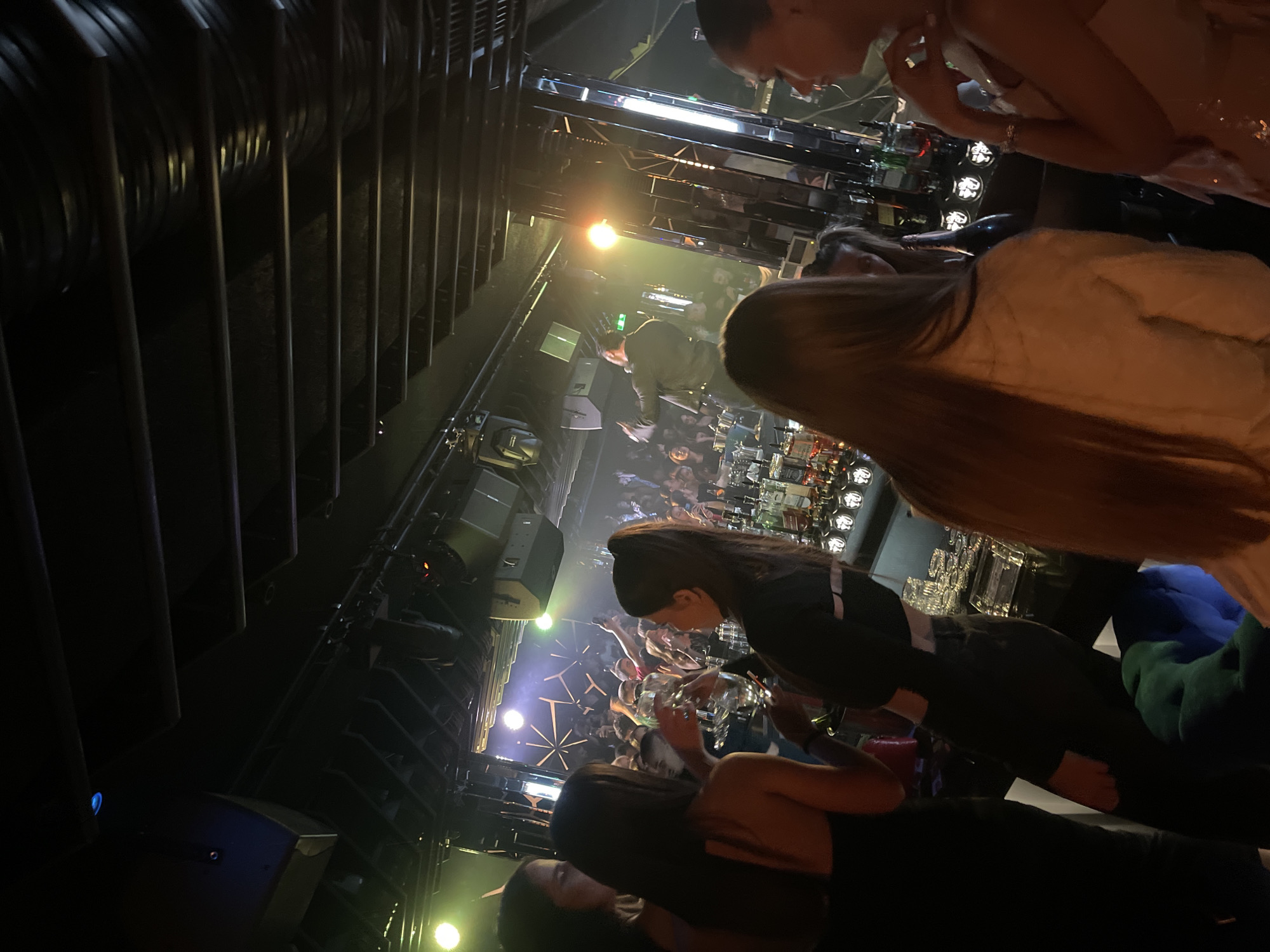 The image depicts the interior of a bar or nightclub in Sofia, Bulgaria. A crowd of people can be seen in the background, illuminated by stage lights and possibly dancing or socializing. Several individuals are positioned closer to the camera, partially obscuring the view of the crowd. These individuals appear to be patrons of the establishment, some holding drinks. The setting suggests a lively and potentially crowded nightlife atmosphere. The foreground shows some structural elements of the building.