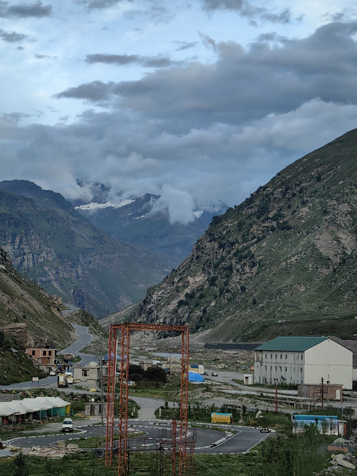 The image depicts a mountainous landscape in India, likely a high-altitude region, given the terrain and the presence of snow-capped peaks in the distance. The scene is dominated by rugged, green-covered mountains that form a valley. A winding road snakes its way through the valley, with several vehicles, including trucks and cars, visible on it. In the foreground, there are some temporary structures, possibly tents or shelters, and a large, orange metal structure, perhaps part of a construction project or an antenna. A multi-story building with a green roof is also present, appearing to be a sort of facility or lodging. The sky is overcast with dramatic clouds, contributing to a somewhat moody atmosphere. The overall impression is one of a remote, mountainous area with some signs of human activity, likely a transit point or a base in a challenging environment.