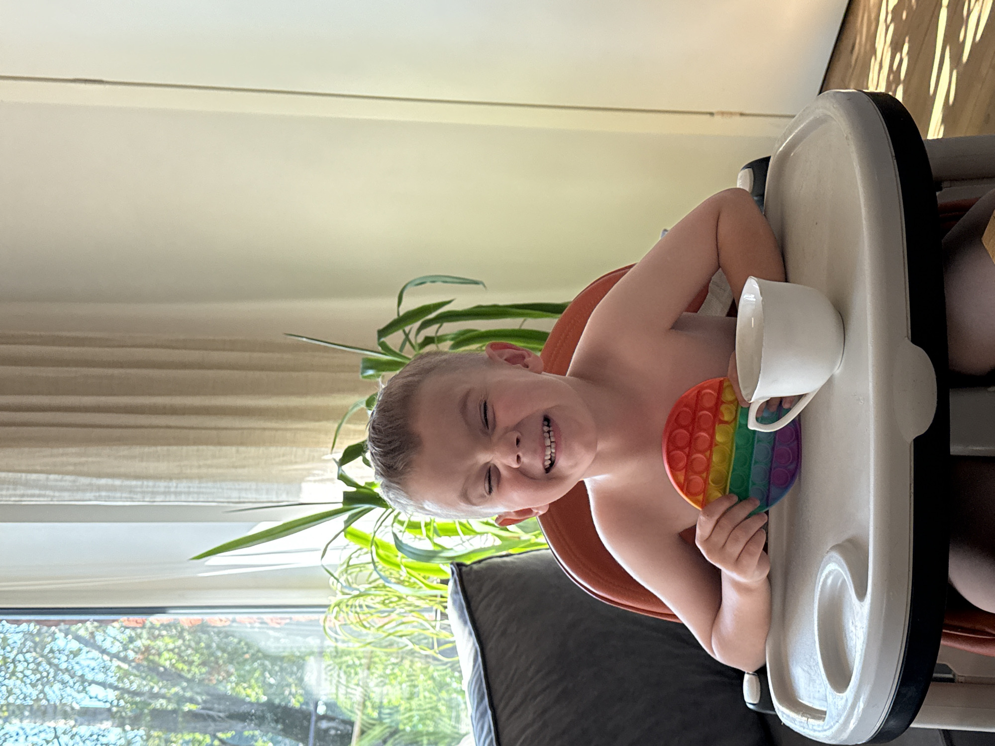 In a well-lit room, a young boy with fair hair and a wide, joyful smile is seated in an orange high chair. He is holding a rainbow-colored pop-it toy in his left hand and a white teacup is visible on the tray in front of him. The background features a large window with sheer curtains, through which sunlight streams in, illuminating indoor plants. The room appears to be a domestic setting, likely a dining area or living space, with a hint of the outdoors visible through the window. The flooring shows dappled sunlight, suggesting a sunny day.