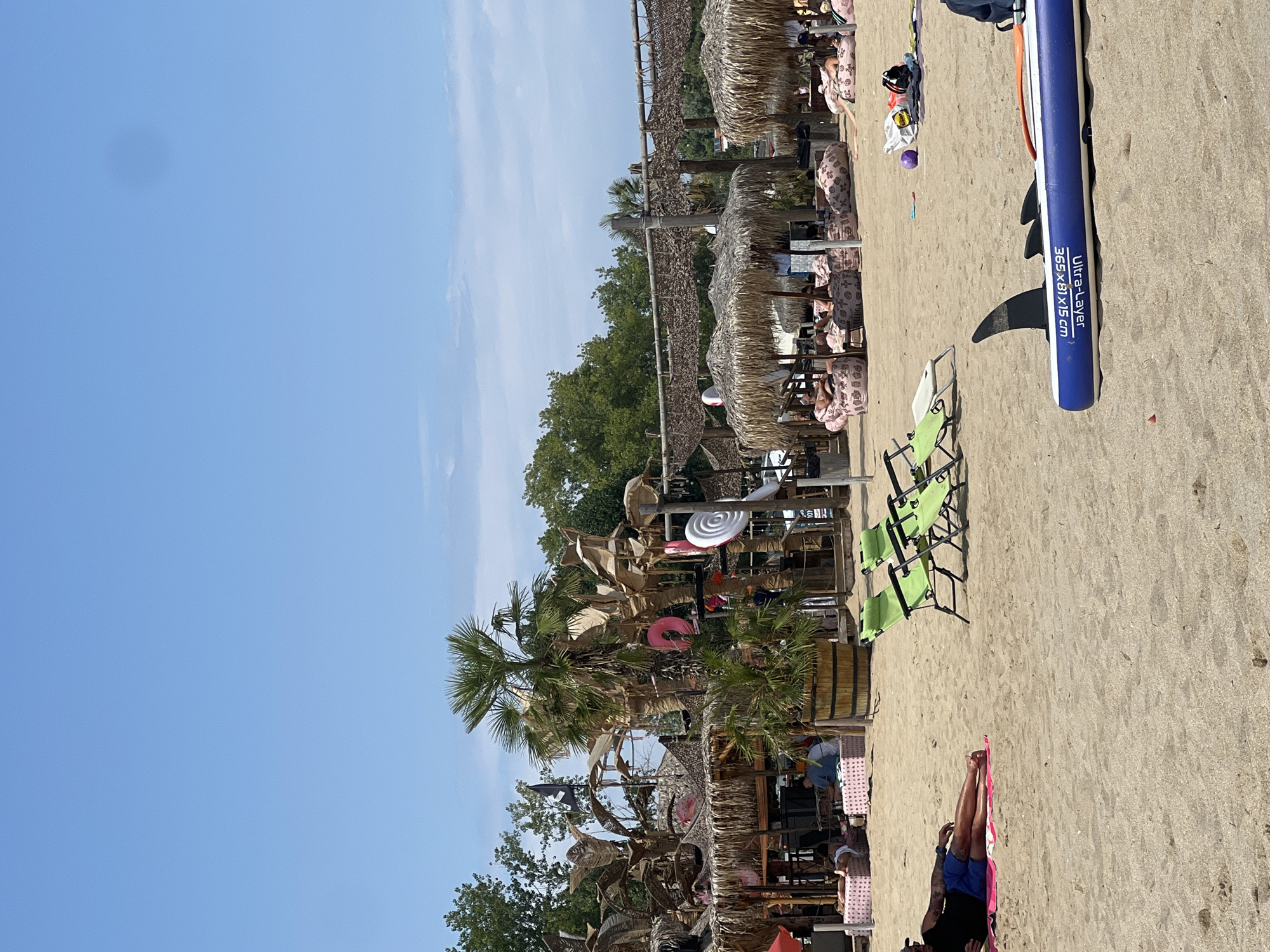 The image depicts a beach scene in Sozopol, Bulgaria, featuring a bar and lounging area. The sandy beach is occupied by several people relaxing on beanbag chairs and loungers under thatched-roof umbrellas. In the foreground, a blue and white paddleboard with a shark fin design lies on the sand, and a person is sunbathing on a mat. The background shows a rustic bar area with palm trees and decorations, suggesting a vibrant atmosphere. The sky is clear blue with a few faint clouds.