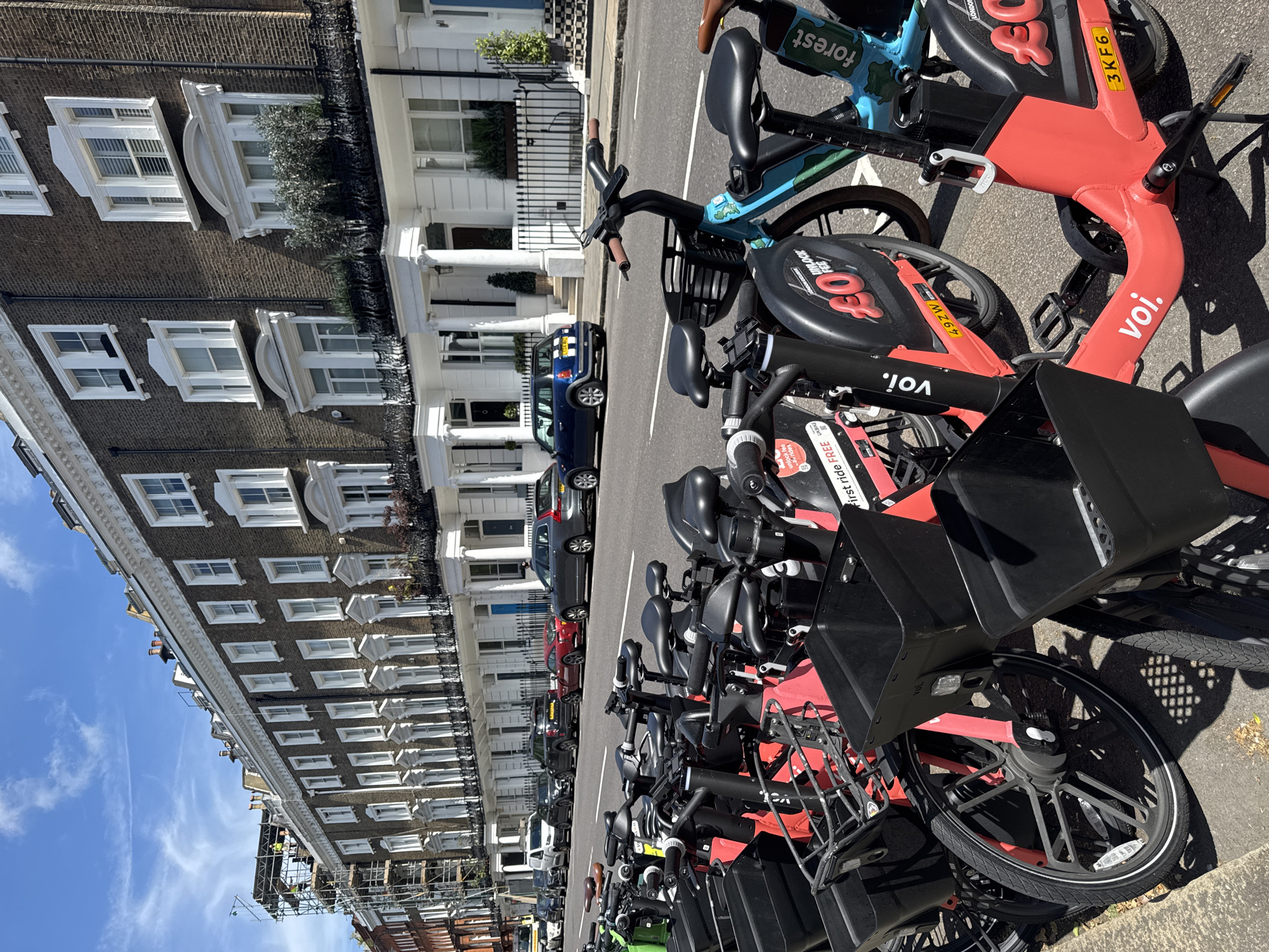 In the city of London, United Kingdom, on a bright, sunny day, a row of electric rental bicycles, primarily red and blue, are parked along a curb on a paved street. The bicycles are modern, with baskets at the front and digital displays indicating rental information, including "voi." and "forest." The street is lined with grand, multi-story Georgian-style townhouses, characterized by their dark brick facades, white window frames, and ornate decorative elements. Some of the houses feature balconies with greenery. In the background, a construction site with scaffolding is visible, with workers present. Several cars, including a red sports car and a dark SUV, are parked on the street in front of the houses. The sky is blue with sparse white clouds. The overall scene depicts a typical London streetscape with a mix of residential architecture, modern transportation, and a hint of ongoing urban development.