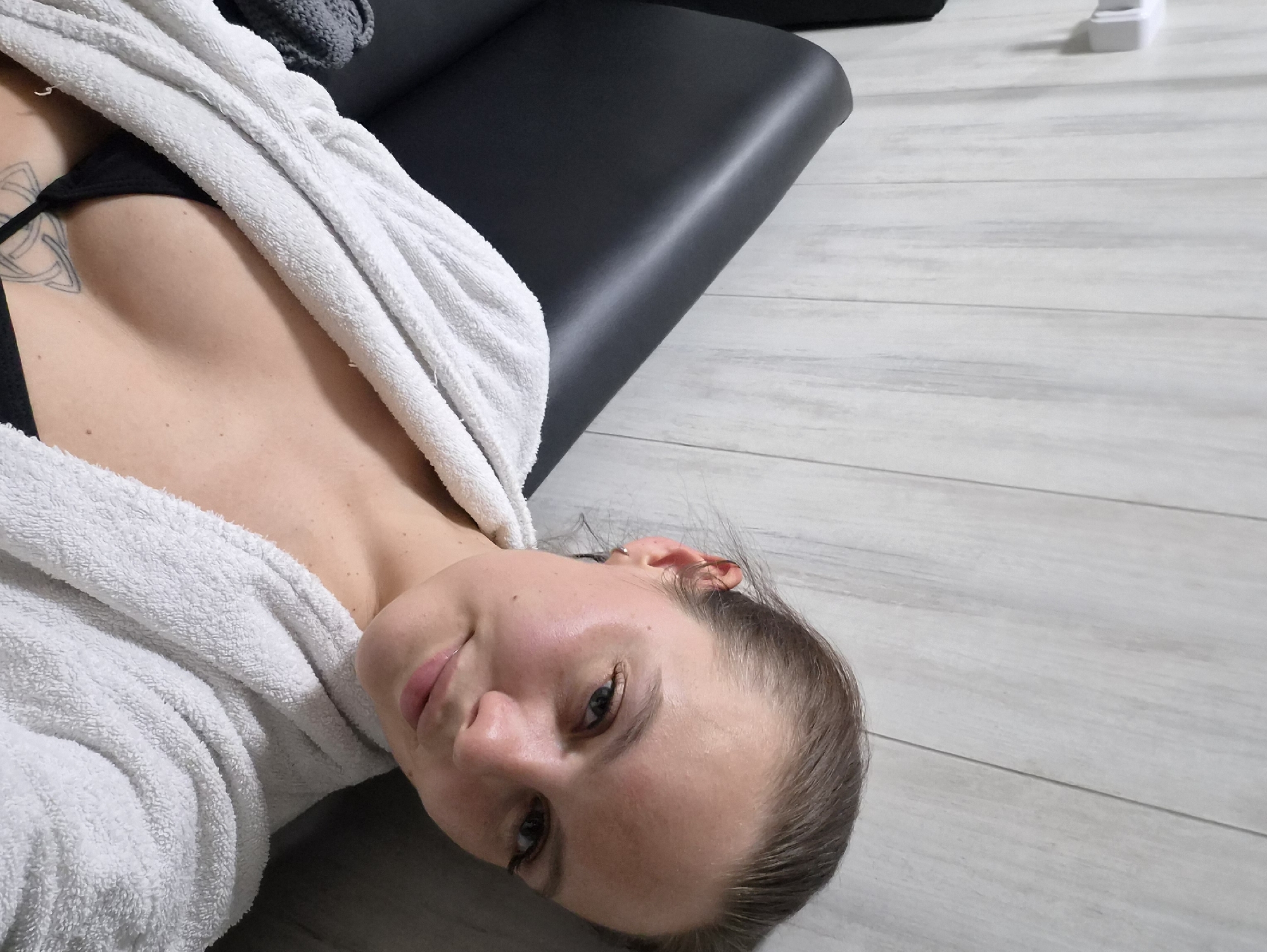 A fair-skinned woman lies on a light-grey, plank-patterned floor. She is wearing a black top and a white robe, which is draped across her torso. A black object, possibly a piece of furniture, is partially visible behind her. The location is Chaikovsky, Russia, but the specific location within the city is not discernible from the image.