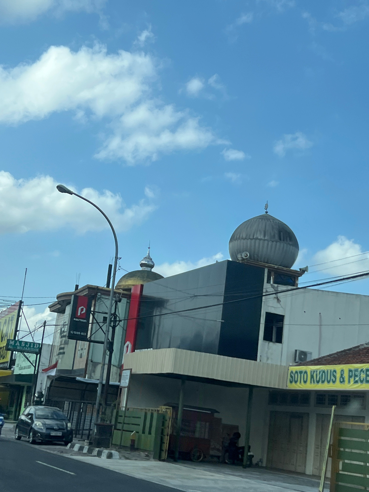 The image captures a street scene in Magelang, Indonesia, on a bright, partly cloudy day. Dominating the mid-ground is a modern building with a prominent dark gray facade and a large, ribbed, gray dome on its roof, suggesting it is a mosque or a religious building. The dome has speakers mounted on its sides. To the left of this building, a street lamp with a curved arm extends into the frame, and a red and black sign with a white "P" logo and the word "POLYTRON" is visible on another building. A sign in green with white text reads "MASJIDI AL-MUJAHIRIN". In the foreground, a dark-colored car is parked on the side of the road, adjacent to a sidewalk with black and white striped curb markings. Further to the right, a yellow banner with green text announces "SOTO KUDUS & PECE". In the background, a few people are visible, including one person sitting on a motorcycle next to a food cart. The sky is a vibrant blue with scattered white clouds.