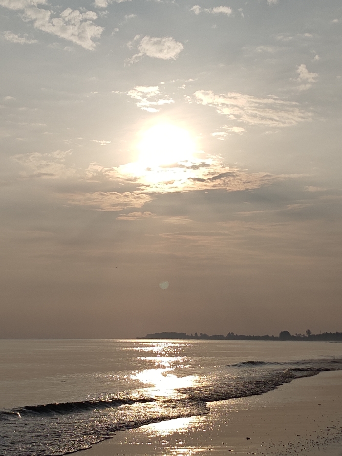 The image depicts a coastal scene in Keude Trienggadeng, Indonesia, observed during either sunrise or sunset. The sun, a bright, diffuse orb, is positioned high in the upper-center sky, partially obscured by thin, scattered clouds, casting a strong glare. The sky exhibits a pale orange-yellow hue near the horizon, transitioning to a light blue-grey higher up, with wispy cloud formations visible throughout. A solitary dark speck, possibly a bird, is visible in the upper-middle portion of the sky. The foreground features a sandy beach, wet from receding waves, with small dark debris scattered on its surface. Ocean waves are breaking gently along the shoreline, creating a foamy edge. The water surface reflects the sun's light in a prominent, shimmering path that extends from the horizon towards the viewer. In the mid-ground, a dark, low-lying landmass or distant coastline is visible, silhouetted against the bright sky, featuring indistinct tree-like forms. No individuals or active interactions are present in the frame.
