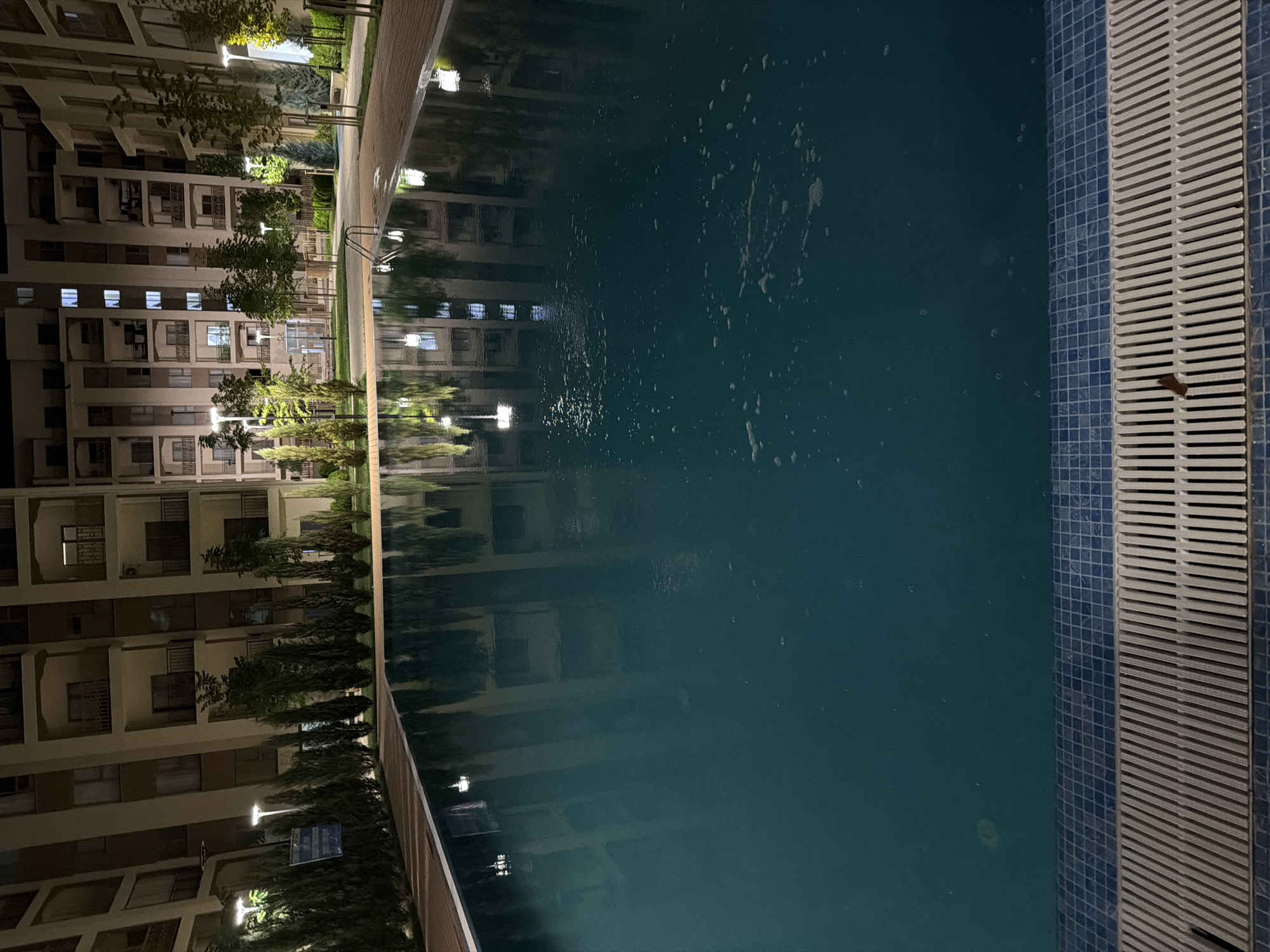 A nighttime exterior view shows a swimming pool adjacent to apartment buildings in Chinarkent, Uzbekistan. The pool water is dark blue and reflects the lights of the surrounding buildings and trees. A metal ladder is partially submerged in the pool. The pool area is bordered by a tiled edge and a grated drain. Behind the pool, tall, slender trees and smaller bushes line a pathway. In the background, multi-story apartment buildings with illuminated windows and balconies are visible. The scene is lit by streetlights and lights from the buildings.