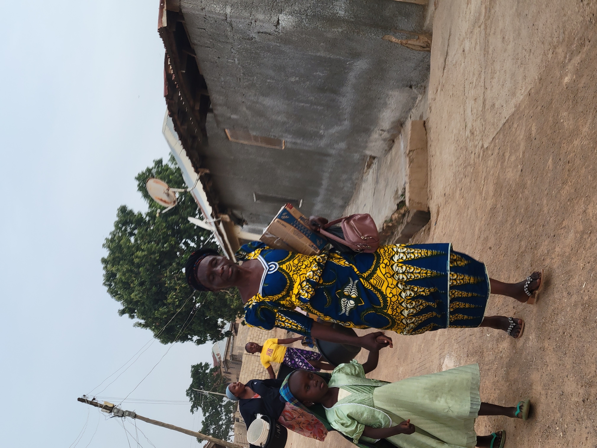 A woman and a young girl are walking hand-in-hand on a dirt path in Jos, Nigeria. The woman wears a blue and yellow patterned dress, black and white sandals, and carries a brown bag and a cardboard box on her left shoulder. The young girl wears a light green dress and green sandals. Other adults and children are walking behind them. Behind them is a rough concrete building with a corrugated metal roof and a satellite dish. A utility pole with wires is visible in the distance on the left. The sky is overcast.