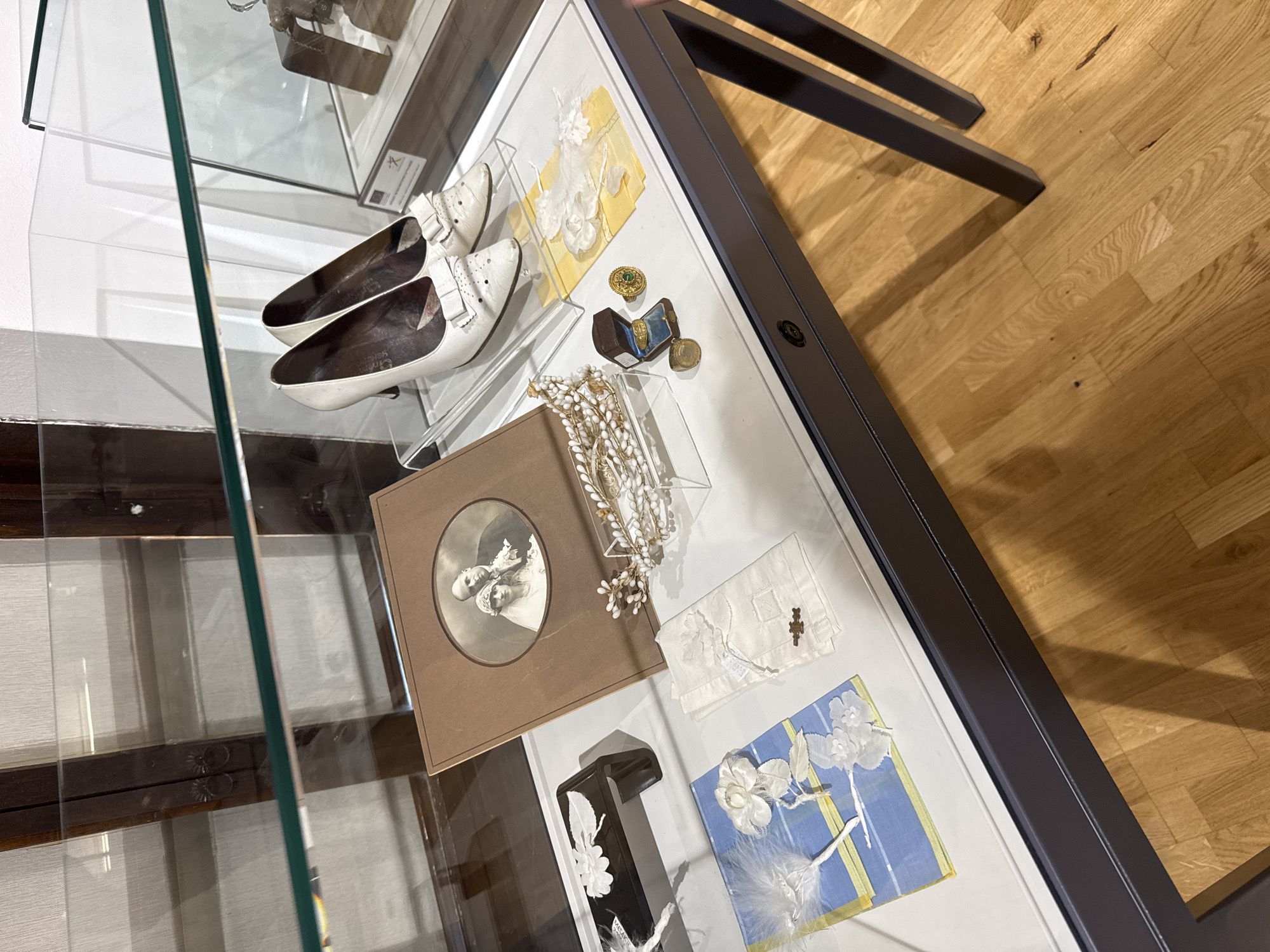 This image shows a display case in the Museum of Humor and Satire in Gabrovo, Bulgaria. The display contains several items, likely related to a wedding or formal occasion. In the foreground, on a white surface, are two white high-heeled shoes with bows. To the left of the shoes is a framed photograph of a couple, appearing to be a wedding portrait. Next to the photograph, to the left, is a small black rectangular object with a white floral decoration. Further to the left is a folded blue and yellow textile item with white floral embellishments. To the right of the framed photograph is a clear box containing a delicate, beaded headpiece or tiara. Below this, there are several rings, including one with a green gemstone, and a small open ring box. To the right of the headpiece is a folded white linen item, possibly a handkerchief, with lace trim and a small brooch. Behind these items, the display case exhibits a dark wooden frame. The floor visible beneath the display case is made of polished light-colored wood planks.