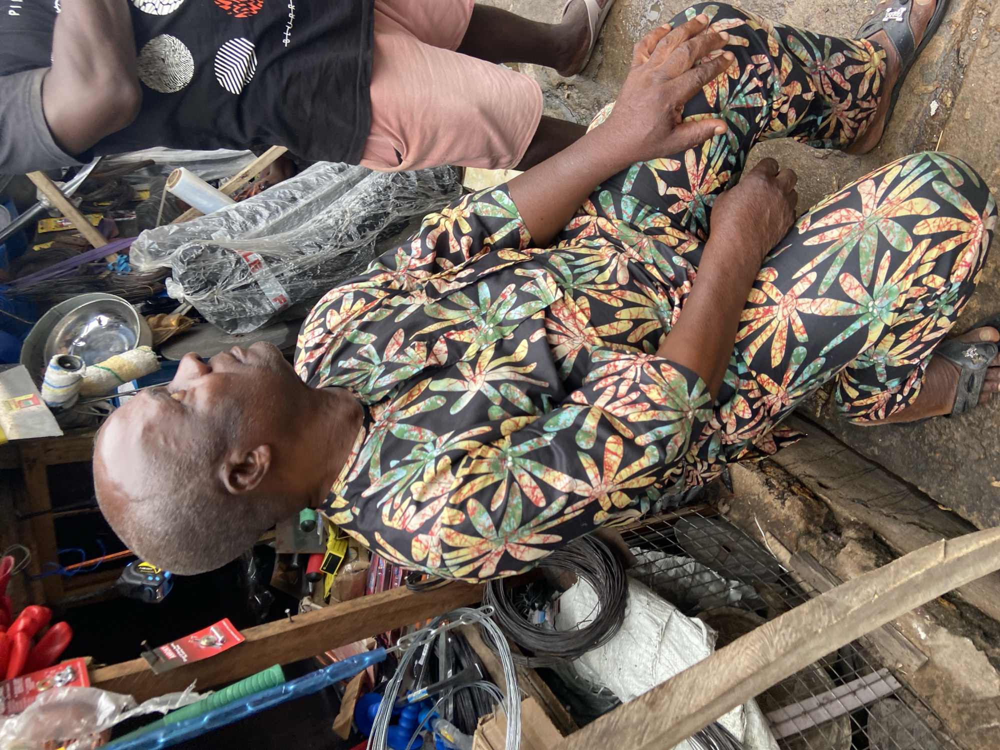 An elderly Black man with short gray hair and brown skin kneels in an outdoor market setting. He wears a black shirt with a vibrant floral pattern in shades of green, yellow, red, and blue, and matching black pants. His right hand rests on his right knee, and his left hand is also on his knee. He is looking to his right, with his head tilted upward.
The environment appears to be a stall or shop, filled with various items. To the left of the man are coiled wires and other metal goods. Behind him, a variety of objects are visible, including tools, plastic-wrapped items, and other market goods. The ground is wet and appears to be made of concrete or similar material. In the background, another person wearing a black t-shirt with a graphic print and pink shorts stands, partially visible. The overall scene suggests a busy marketplace.
The man is identified as a resident of Ogbunike, Nigeria, a town in the Anambra State of Nigeria known for its caves. This specific location within Ogbunike is an outdoor market stall.