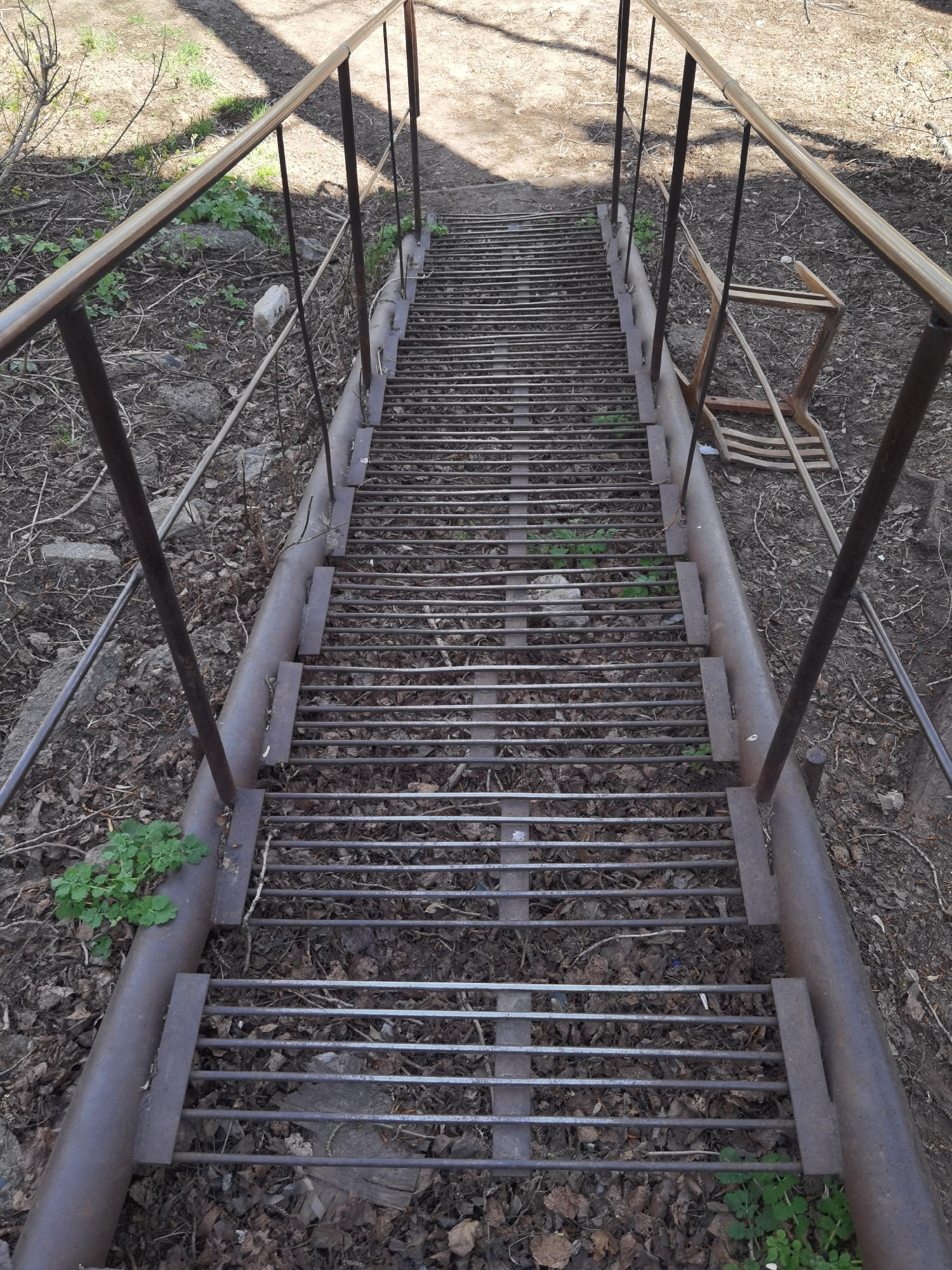The image shows a long metal staircase leading uphill. The staircase is constructed with metal pipes for the sides and railings, and metal rods forming the steps. The steps are covered in dried leaves and debris. Vegetation, including grass and small plants, grows through the steps and along the sides of the staircase. A broken wooden chair is visible to the right of the staircase, partially obscured by the railing. The surrounding area consists of bare earth and fallen leaves, with some patches of grass. No people are visible in the frame. While the location is mentioned, I am unable to verify the city in which this picture was taken.