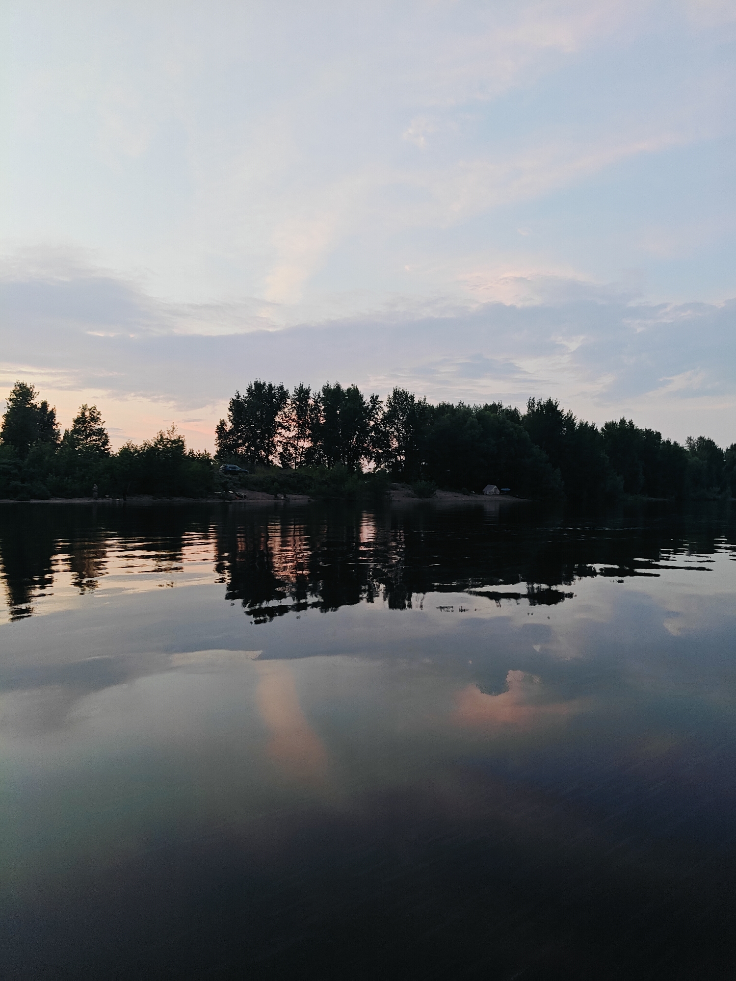 The image depicts a tranquil river scene at dusk. The sky is a soft gradient of pale blue and white, with wispy clouds tinged with pink from the setting sun. A line of dark green trees forms a dense canopy along the riverbank, with a sandy stretch visible in places. The trees are reflected clearly in the calm, dark water, creating a mirror image that blurs into the twilight. The water's surface is disturbed by gentle ripples, adding texture to the reflection. There are no visible people or vehicles in the image, emphasizing the solitude and natural beauty of the location. The scene is likely situated in Noviy, Russia, as indicated by the typical landscape of forested riverbanks common in the region during this time of year.