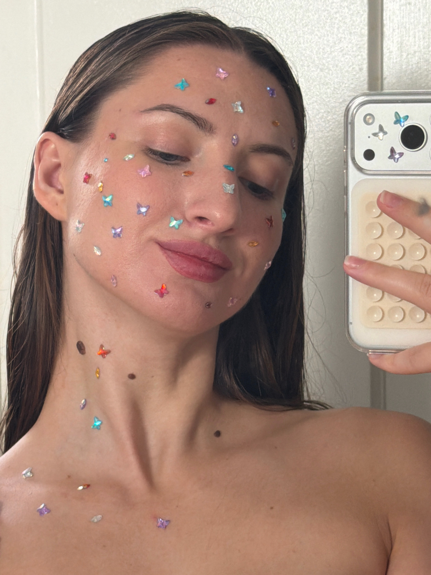 A woman with dark hair, adorned with colorful butterfly stickers on her face, neck, and décolletage, holds a smartphone in her right hand, capturing a selfie. The stickers are scattered across her skin, some appearing to be translucent and iridescent. The woman's eyes are closed, and her lips are slightly parted. She is positioned in an indoor setting, with a plain white wall visible in the background. The smartphone case also features butterfly stickers.