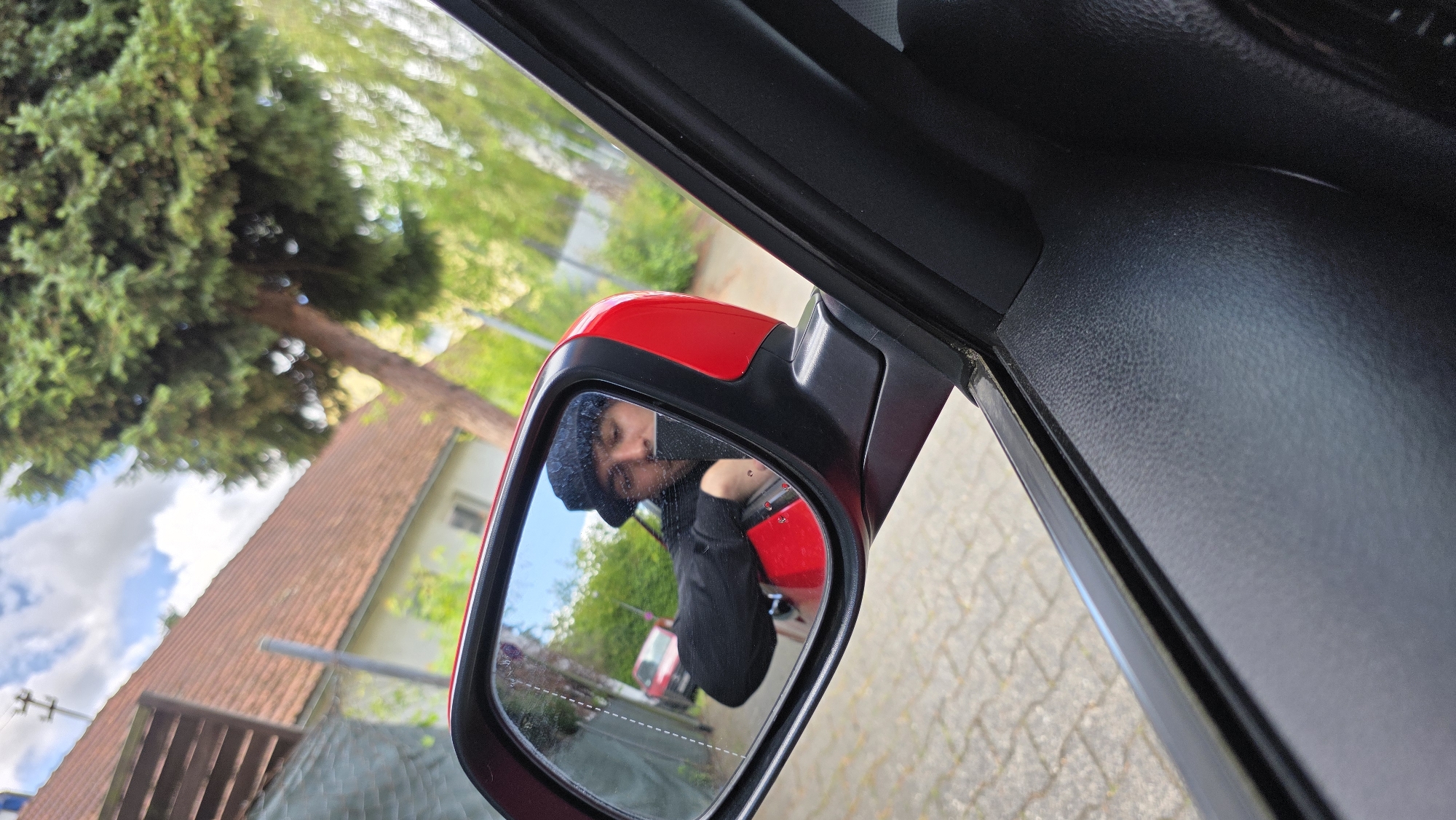In this image, a man wearing a dark hat and a black sweater is reflected in the side-view mirror of a red car. He is leaning out of the car window, holding what appears to be a phone. The reflection shows a residential street with houses and trees in the background. The car is parked on a paved surface.