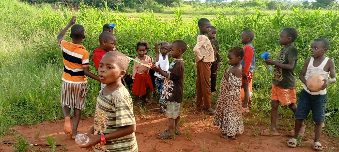 The image displays approximately 14 children, appearing to be between 3 and 12 years of age, gathered on a reddish-brown dirt path in Ngalula, Democratic Republic of the Congo. The immediate foreground shows a child in a striped t-shirt holding a clear plastic bottle, looking towards the left with their mouth slightly open. To their right and slightly behind, another child in an orange, white, and black striped shirt faces away with their right arm raised.
Several other children are distributed across the scene. A child in a white tank top and camouflage shorts holds a colorful toy constructed from plastic scraps. Another child in a dark green t-shirt and orange shorts holds a blue funnel-like object. Adjacent to them, a child in a white tank top and dark blue shorts holds a light brown spherical object resembling a deflated football. One young child wears a leopard-print dress. Most children are barefoot, while some wear sandals.
The environment consists of an unpaved dirt ground with sparse green sprouts. The background is dominated by dense, tall green vegetation, indicative of overgrown fields or natural foliage. A faint, rudimentary metal frame, possibly a goalpost, is visible through the distant vegetation in the upper left quadrant. The scene is illuminated by natural daylight.