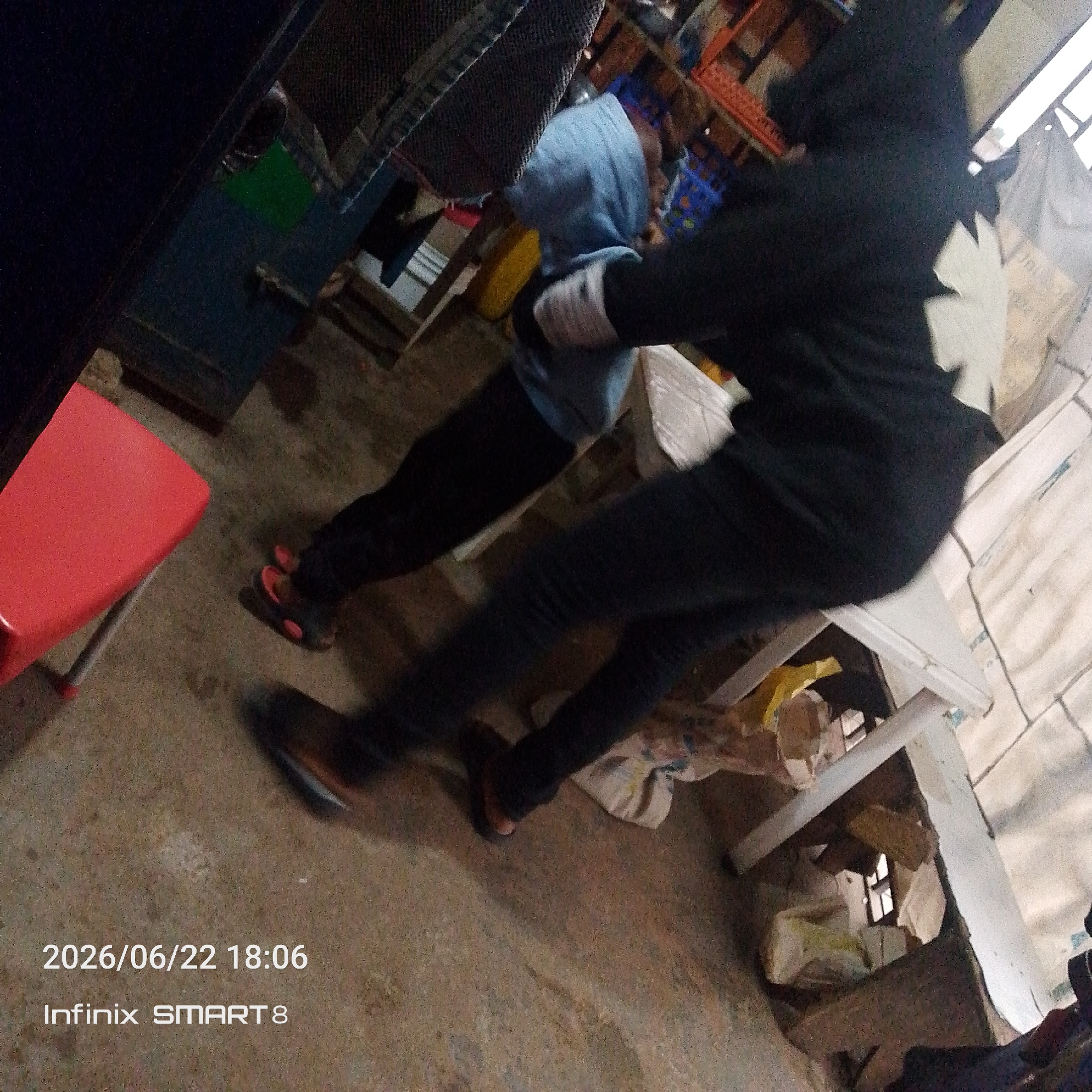 In this image, two individuals are present in what appears to be an indoor, cluttered setting. One person is wearing a blue hooded sweatshirt and dark pants. The other person is dressed in a black hooded outfit with white detailing on the back, possibly a symbol or design. They are positioned facing each other, and their bodies suggest some form of interaction or movement, though the exact activity is unclear due to the motion blur.
In the foreground, a bright red plastic stool is visible on the left. The ground appears to be a rough, textured surface. The background is filled with various objects, including what looks like shelving units stacked with items, some wrapped in plastic or cloth. There's a general sense of disarray, with boxes and other materials scattered around. A white plastic bag is hanging on the right side of the frame.
The image contains visible text overlay in the bottom left corner: "2026/06/22 18:06" and "Infinix SMART 8". This indicates the date and time the photo was taken, as well as the device used. The lighting suggests it might be late afternoon or early evening, given the "18:06" timestamp. The environment doesn't provide clear clues about the weather or a specific geographic location, beyond being indoors. The mood of the scene is difficult to ascertain due to the motion blur and the general nature of the setting.