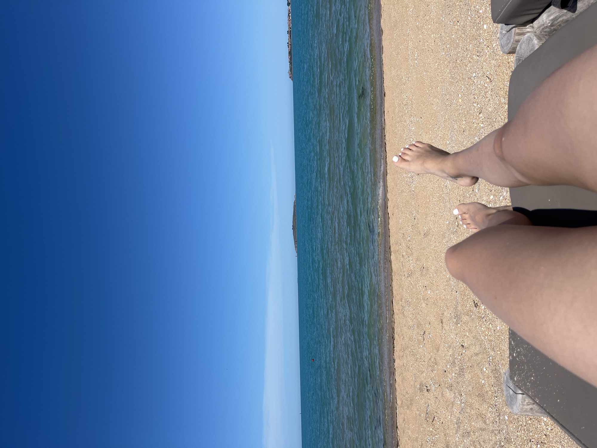 The image shows a first-person perspective of someone relaxing on a beach chair on a sandy beach. The person's legs and feet, with white-painted toenails, are visible in the foreground. Beyond the beach, the turquoise sea stretches out to the horizon under a clear blue sky. In the distance, there are a couple of small islands or peninsulas, and a red buoy is visible in the water. The scene appears to be a tranquil beach setting, possibly in or near Sozopol, Bulgaria, given the coastal environment and clear skies indicative of a Mediterranean climate. The overall atmosphere is one of leisure and relaxation.