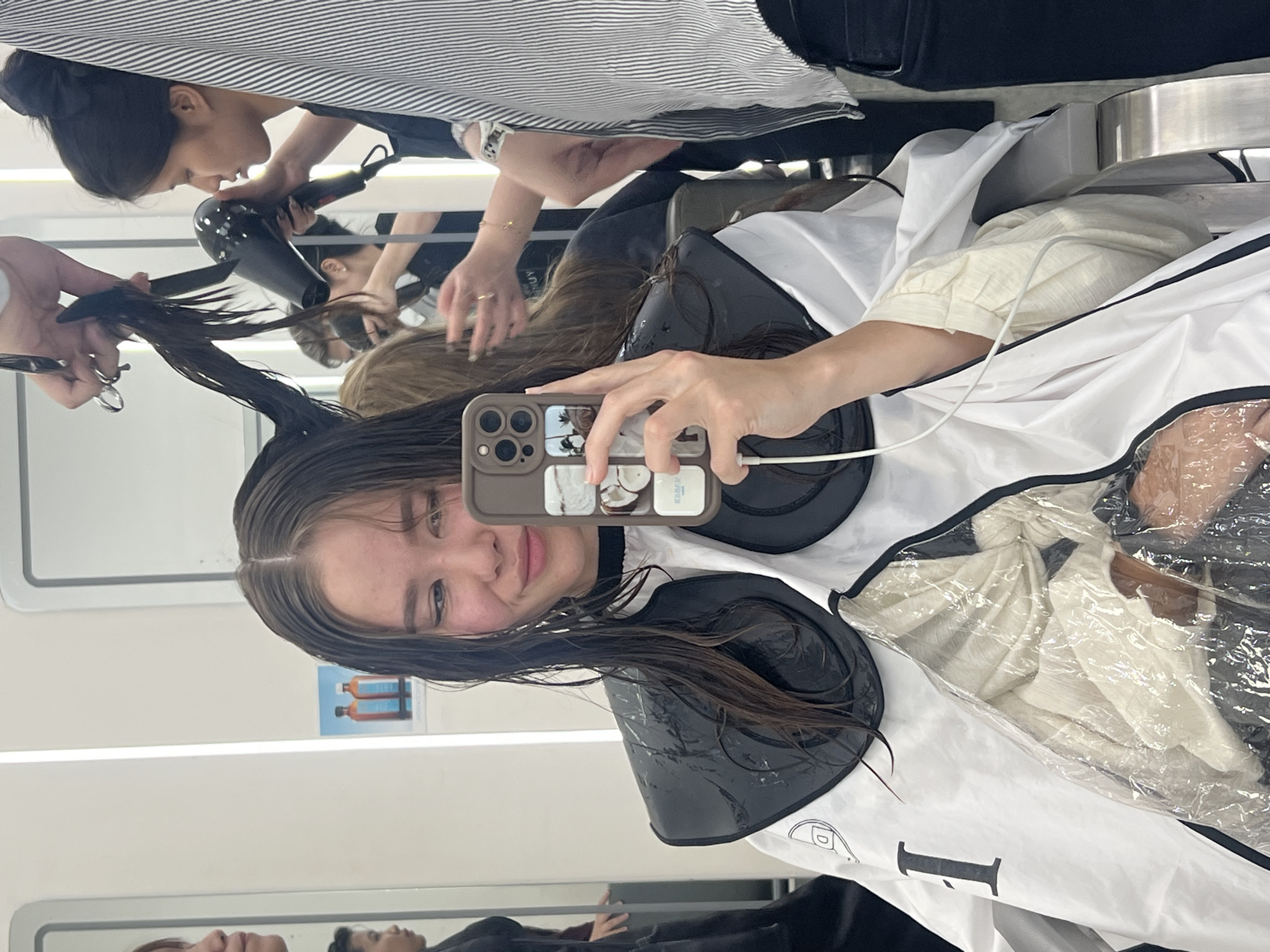 The image displays an interior scene at a hair salon in Nha Trang, Vietnam. A young female client is seated, taking a selfie with a tan-cased iPhone held in her right hand. Her wet, dark brown hair is styled with a center part. She wears a white, black-trimmed hair styling cape with transparent plastic sections over the chest, and a black, plastic shoulder protector, both visibly wet with water droplets. A white cable from her phone extends downwards.
Behind the client, a female hair stylist, reflected in the prominent mirror, actively cuts a section of the client's elevated hair with silver shears held in her right hand, while simultaneously drying it with a black hair dryer held in her left. This stylist has dark hair secured in a bun and wears a dark uniform. To the right, another individual, wearing a striped shirt and dark pants, is partially visible, with her reflection further back suggesting she is attending to another client's brown hair.
The salon environment is brightly lit, characterized by white walls, integrated vertical light strips, and large mirrors. A blue poster featuring two amber bottles and the reversed text "BOHASSIN" is reflected on the wall behind the client. A metallic armrest of a salon chair is partially visible on the right.