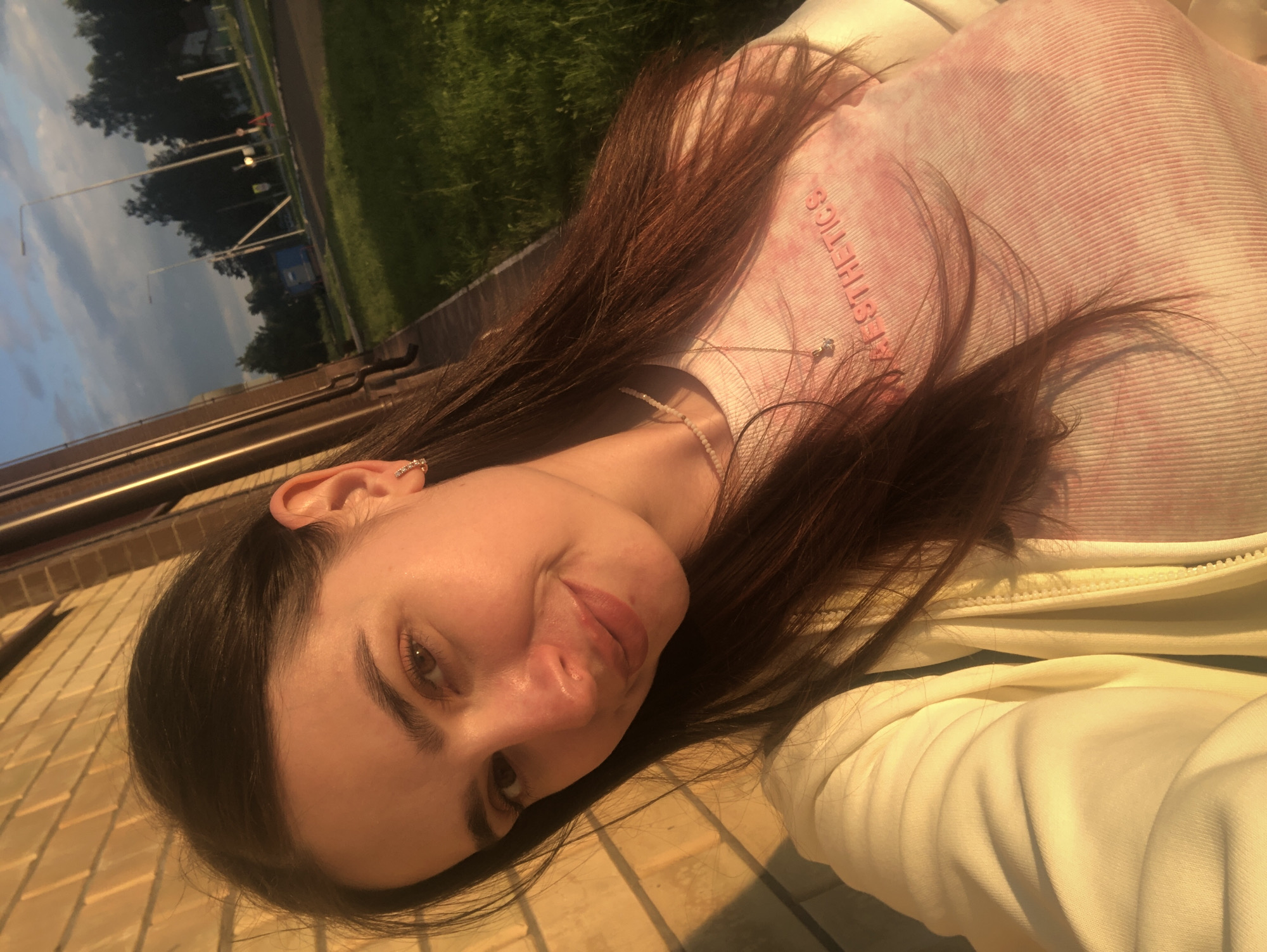 A young woman with long brown hair and a light pink tie-dye shirt, adorned with a delicate necklace, is captured in a selfie. She's wearing a light-colored zip-up jacket. The setting appears to be outdoors in a suburban area, possibly Noviy, Russia, as indicated by the presence of a brick building, grassy areas, trees, and a street with lampposts in the background. The golden hour light casts a warm glow on the scene.