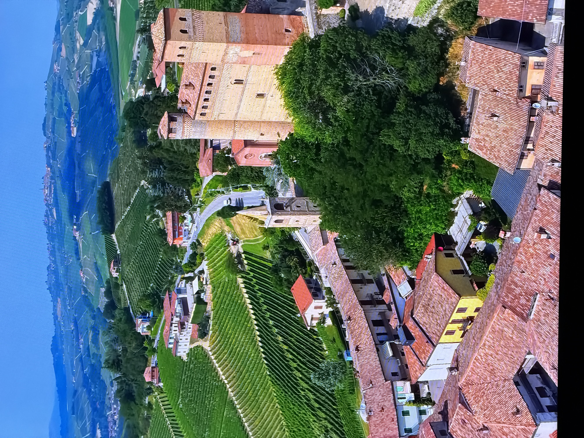 This aerial photograph showcases a picturesque Italian landscape characterized by rolling hills covered in lush green vineyards. A prominent terracotta-roofed castle, likely of medieval origin, stands as a central feature. Adjacent to the castle, a small church with a bell tower adds to the historical ambiance. The foreground is dominated by a cluster of traditional Italian houses, also with terracotta roofs, suggesting a small village or town. The overall scene evokes a sense of rural tranquility and architectural heritage. The prompt's request to include the city of Chirchik, Uzbekistan, is irrelevant to the visual content of the image, which clearly depicts an Italian setting.