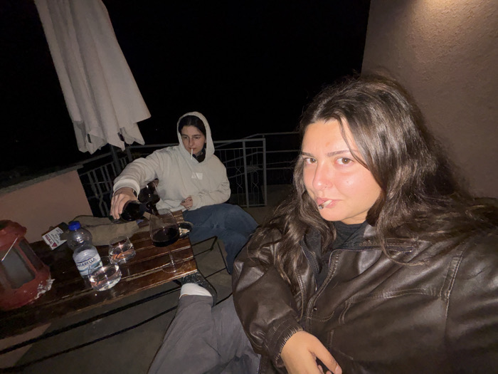 This image depicts a nighttime scene on an outdoor balcony or terrace in Nesso, Italy. Two females are present. In the foreground, a female with long, dark, wavy hair wears a brown leather jacket and is looking directly at the camera, with a metallic object protruding from her lower lip. In the background, a second female, wearing a light-colored hooded sweatshirt and dark jeans, is seated and actively pouring a dark red liquid from a dark bottle into a clear tumbler on a wooden table. A lit cigarette is visible in her mouth. The wooden table holds a red lantern-like object, a small white packet, a clear plastic bottle labeled "FONTARIELLA" (water), and three clear tumblers, two containing liquid and one receiving the poured liquid. An off-white patio umbrella is partially visible in the upper left, and a metal railing is situated behind the table. The background beyond the railing is entirely dark, indicating night. A light-colored wall is visible on the right side of the frame.